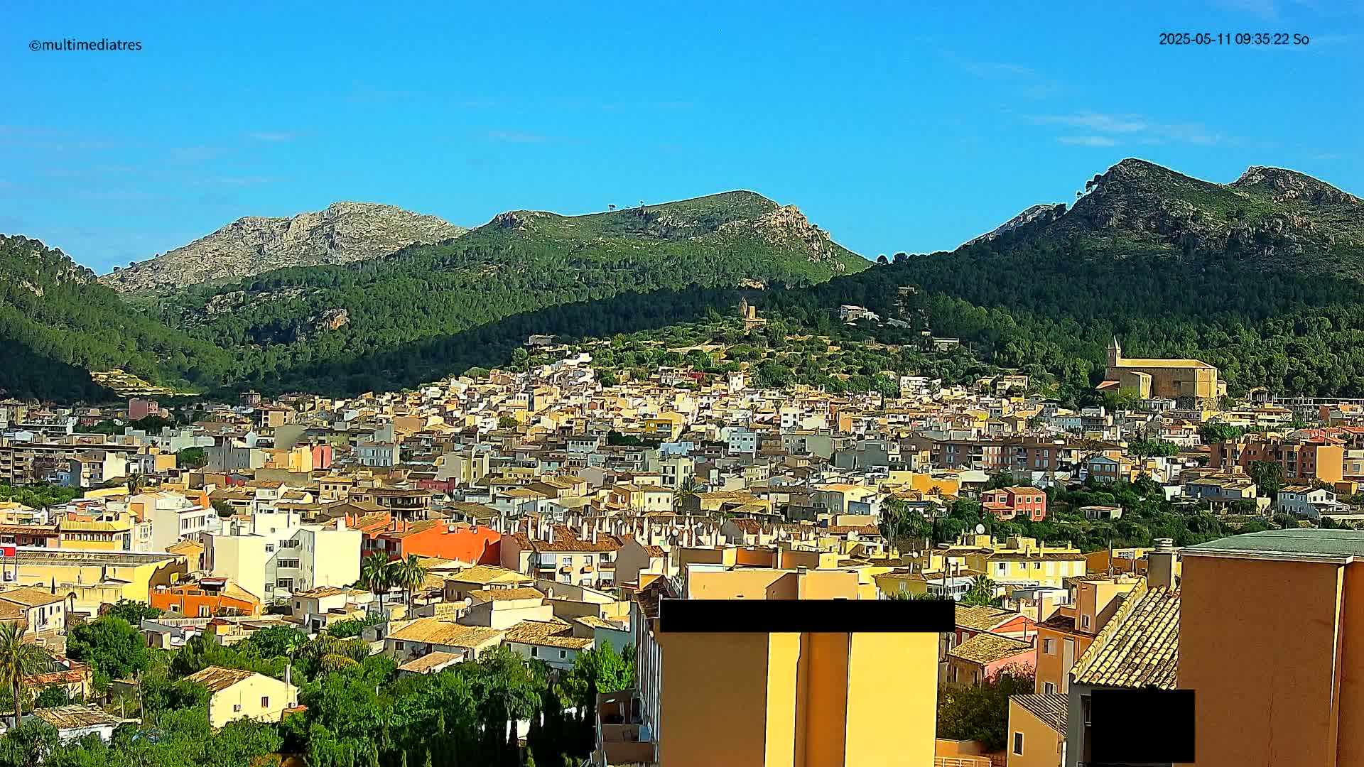 A sprawling town of low-rise buildings nestles at the base of verdant, mountainous terrain under a clear blue sky.