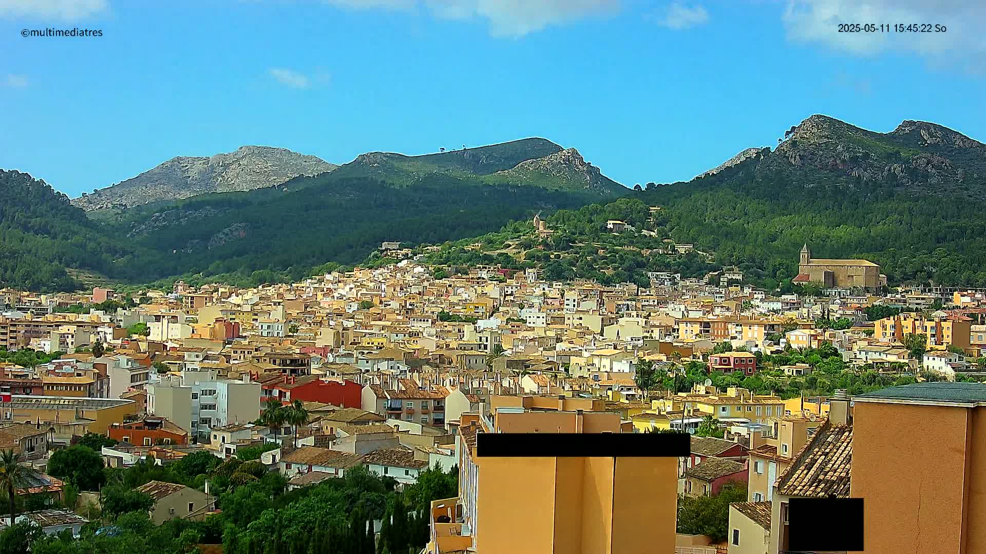 A sprawling town of low-rise buildings nestles at the foot of verdant, rocky mountains under a mostly clear, sunny sky.