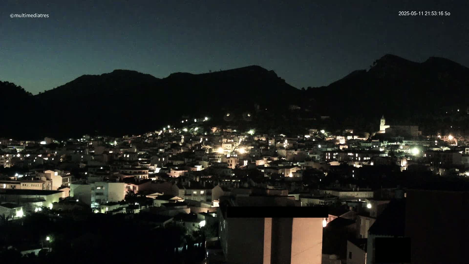 A nighttime view of a town nestled against a dark mountain, illuminated by scattered lights.
