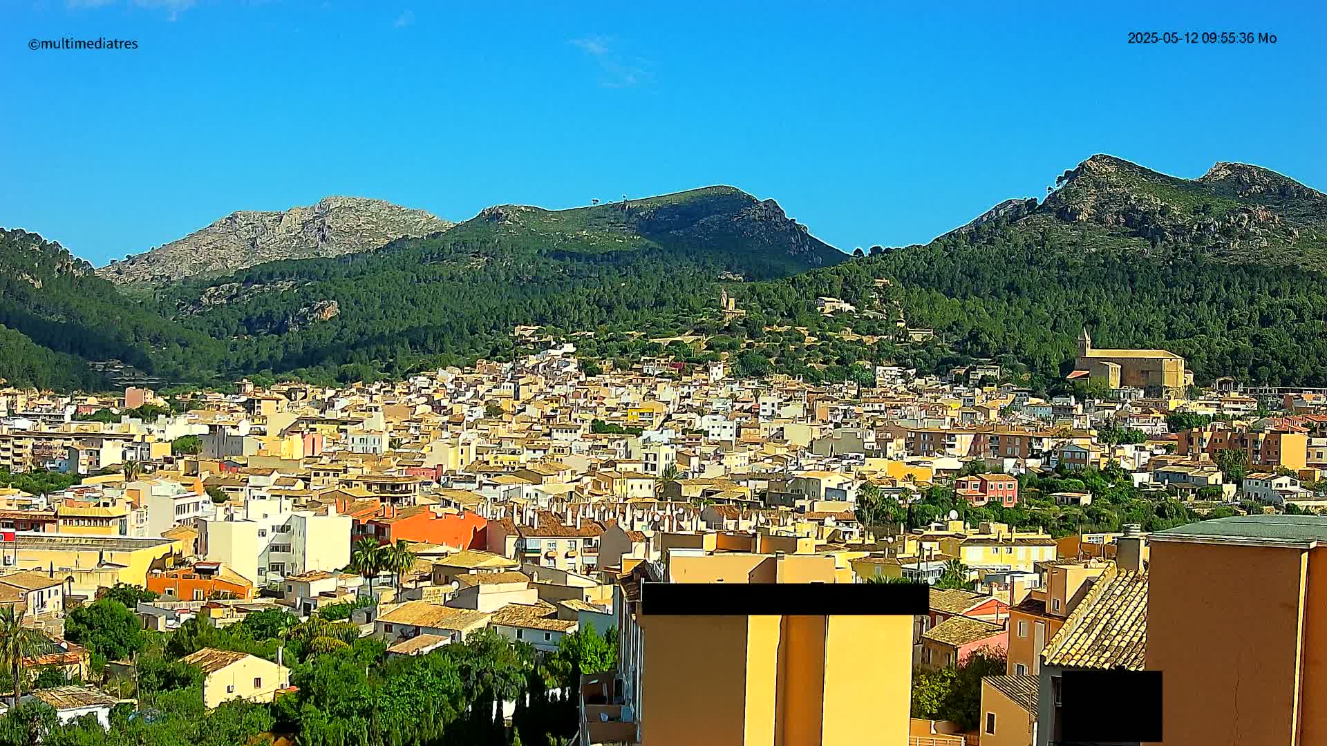 A sprawling town of low-rise buildings nestles at the foot of verdant, rocky mountains under a clear blue sky.