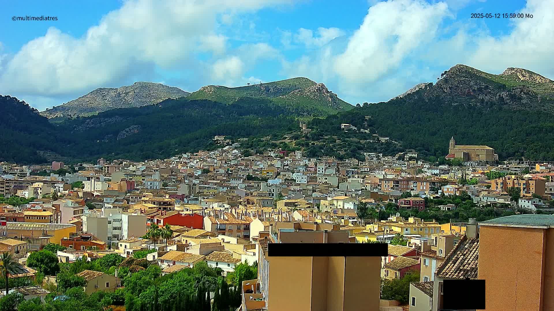A sprawling town of low-rise buildings nestles at the foot of lush, green mountains under a partly cloudy blue sky.
