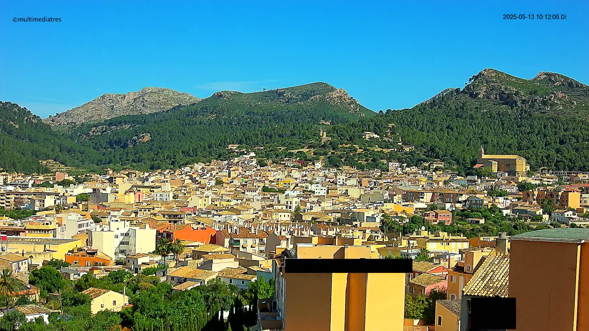 A sprawling town of tan and orange buildings nestled in a valley surrounded by lush green, tree-covered mountains under a clear blue sky.