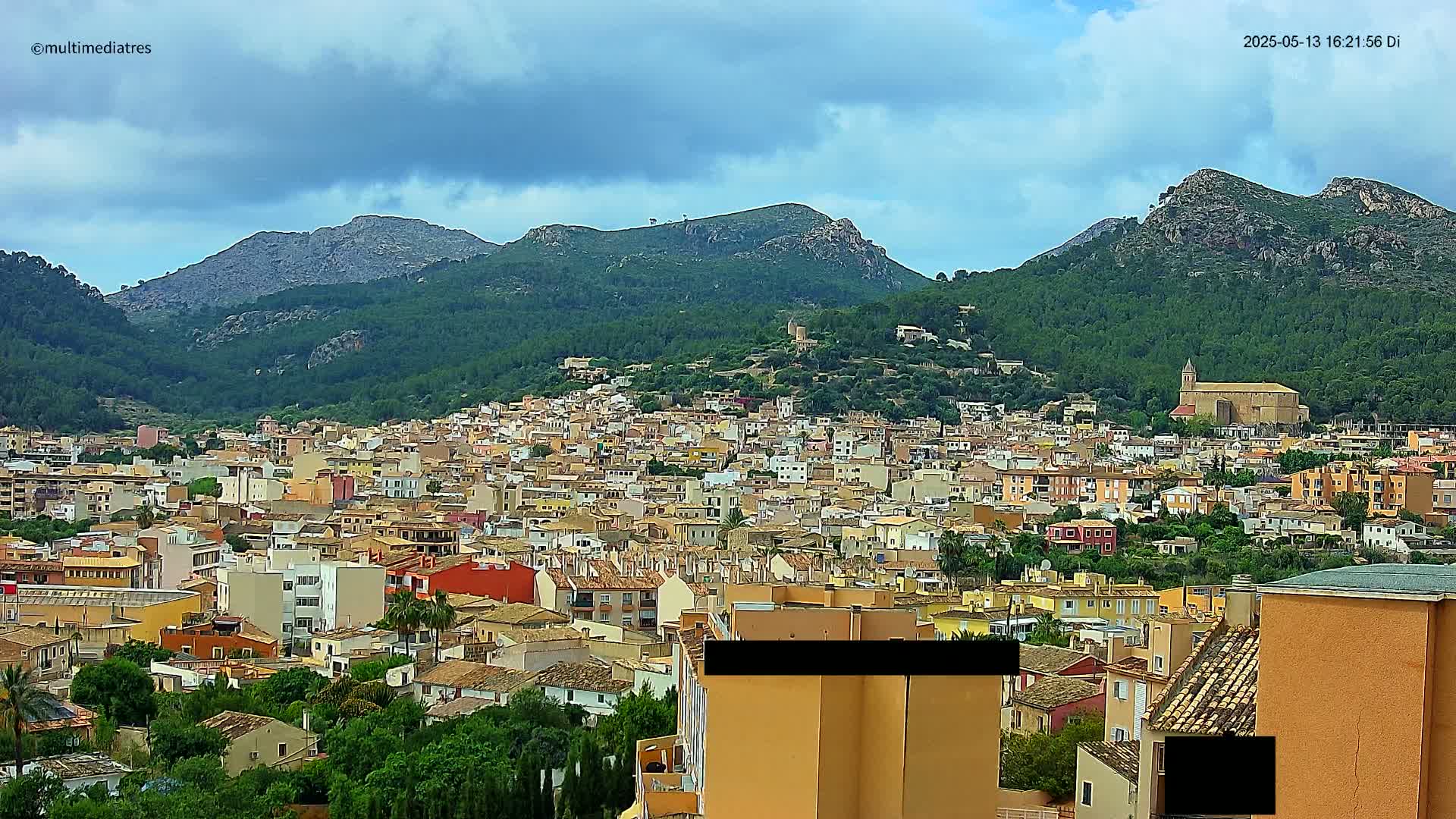 A sprawling town of tan and orange buildings nestles in a valley between verdant, rocky mountains under a partly cloudy sky.