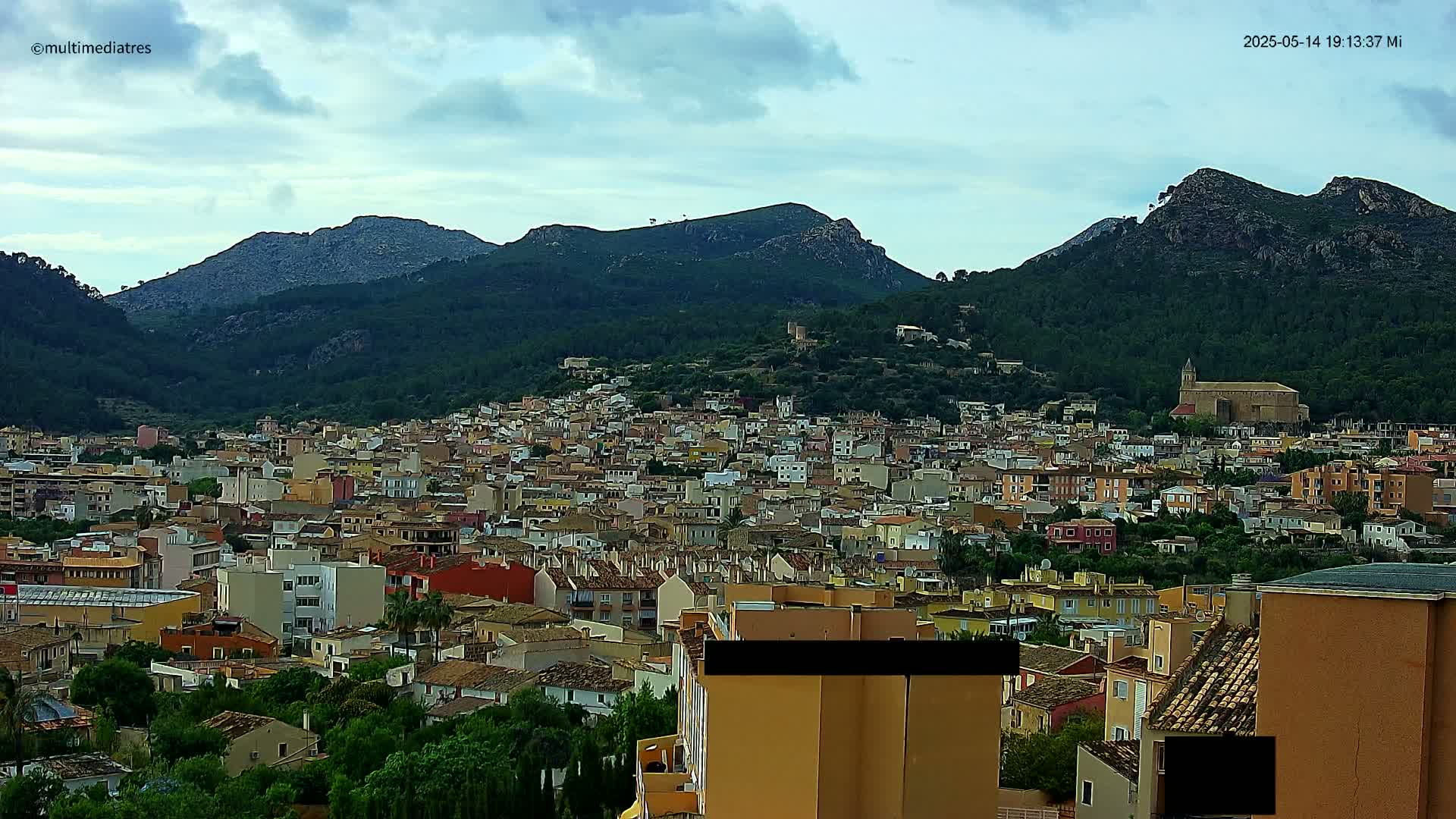 A sprawling town of low-rise buildings nestles at the foot of verdant mountains under a mostly cloudy sky.