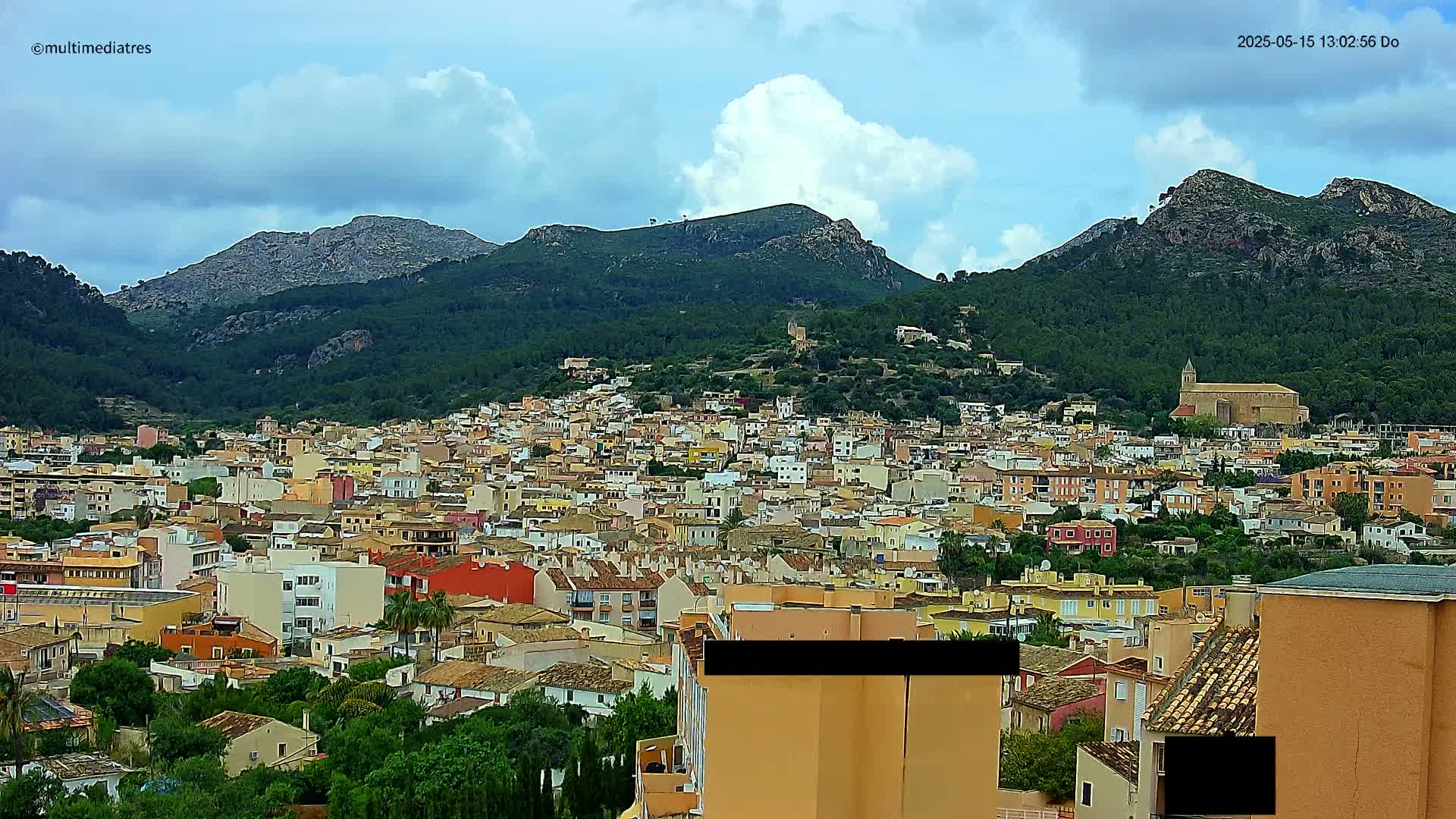 A sprawling town of colorful buildings nestled in a valley between verdant, mountainous hills under a partly cloudy sky.