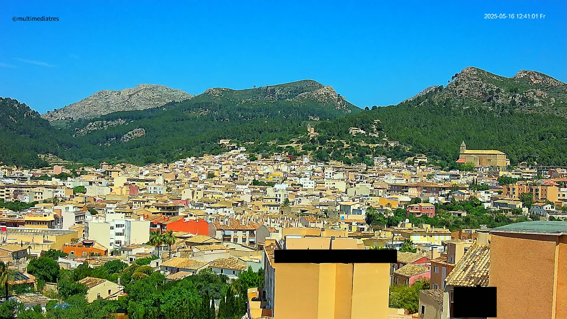 A sprawling town of tan and orange buildings nestles in a lush green valley, backed by rocky, tree-covered mountains under a clear blue sky.