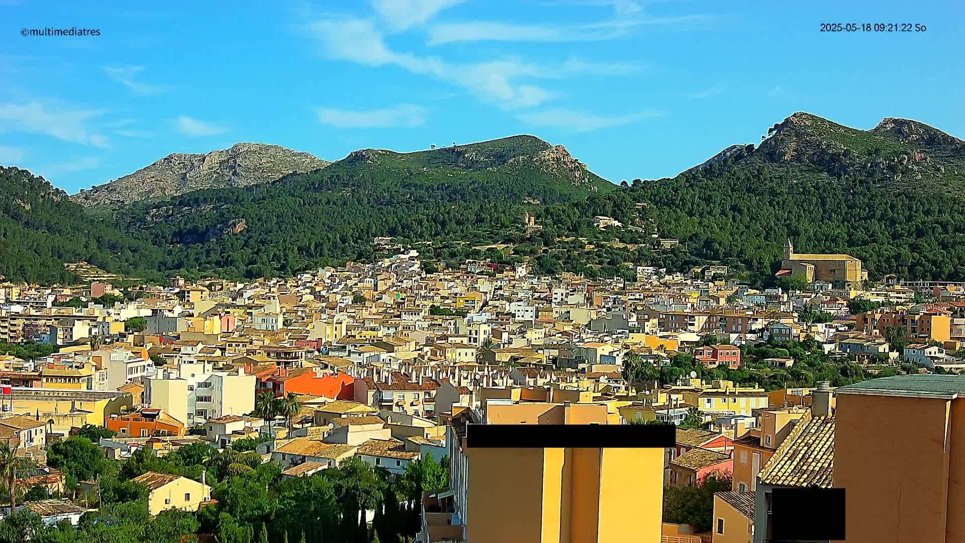 A sprawling town of low-rise buildings nestles at the foot of verdant, rocky mountains under a clear, bright blue sky.