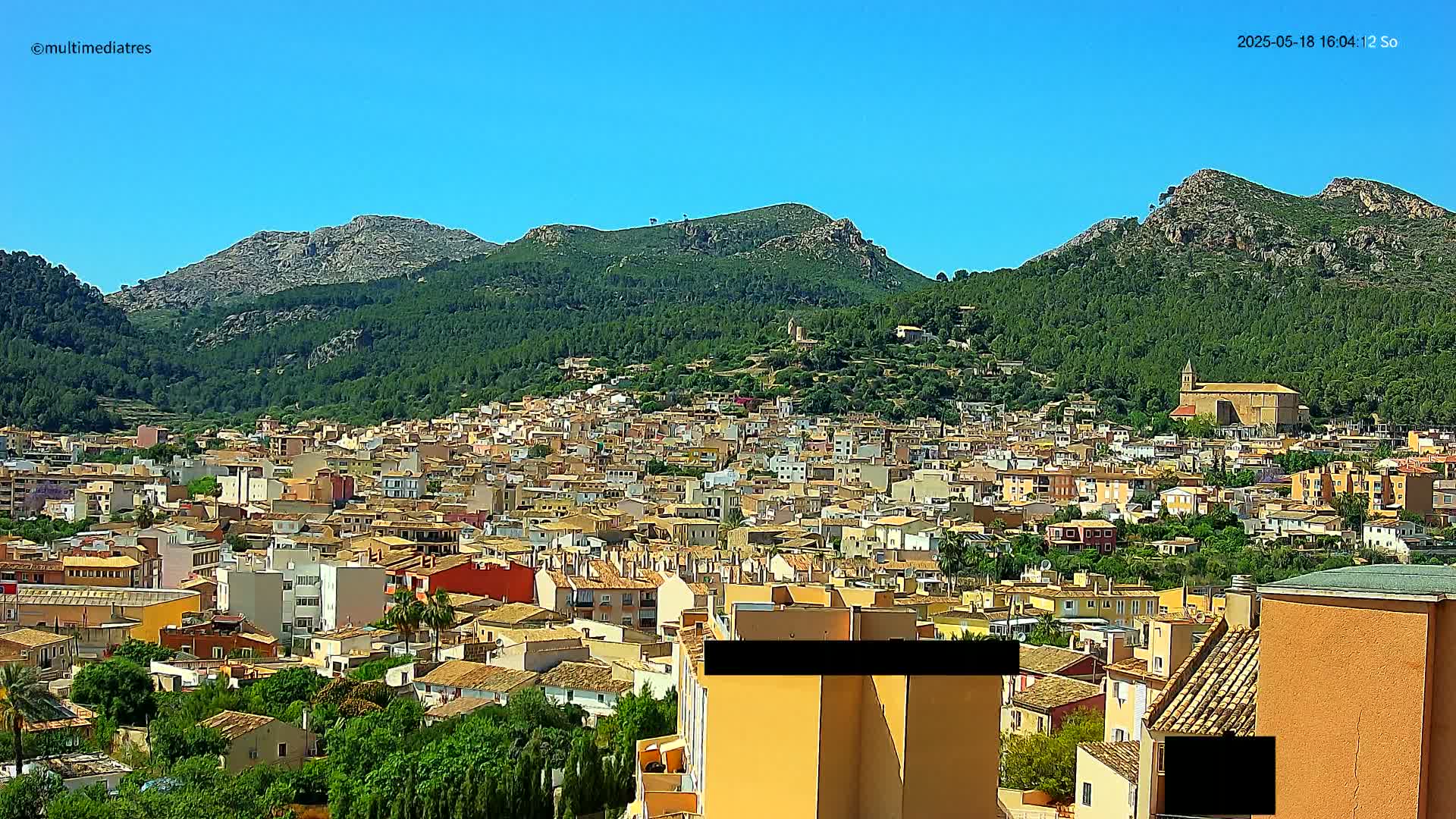 A sprawling town of tan and red-roofed buildings nestled in a valley, surrounded by lush green hills and mountains under a clear blue sky.