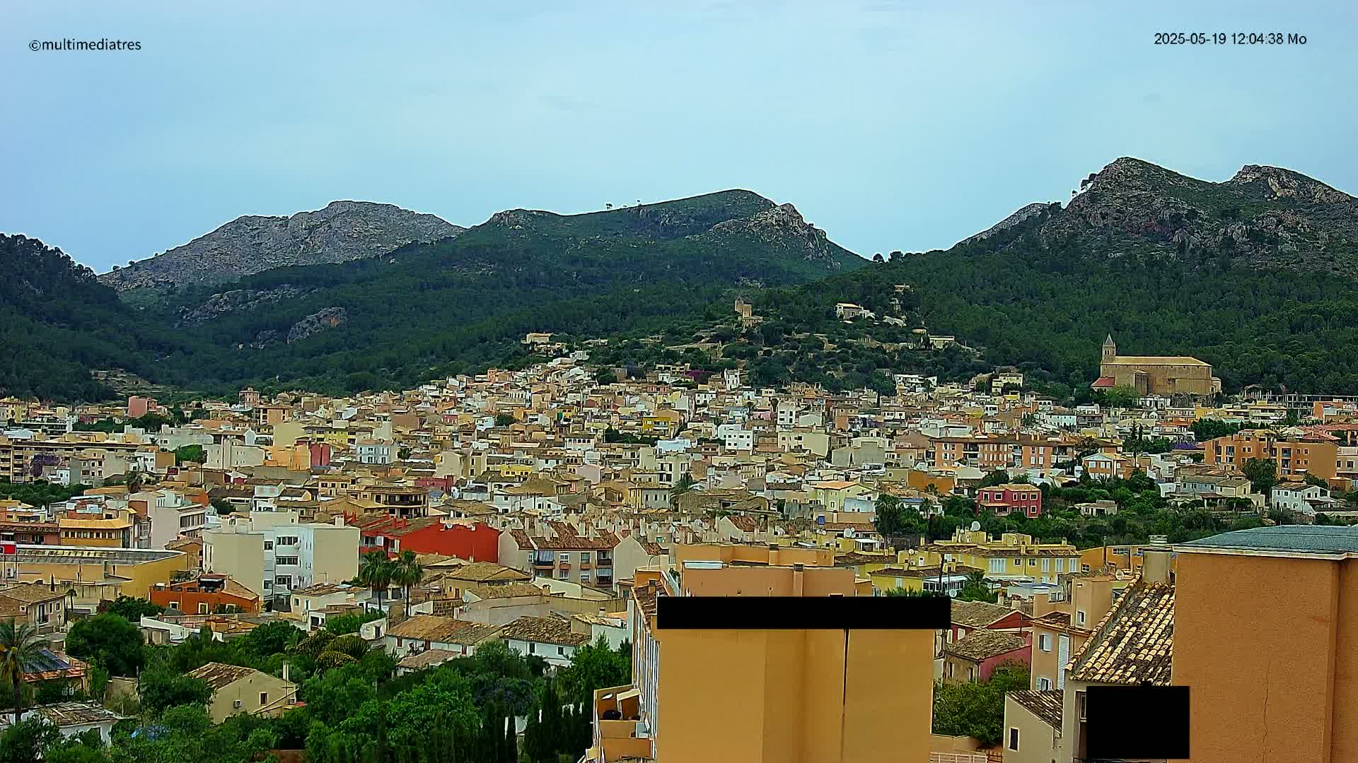 A sprawling town of low-rise buildings nestled in a valley between verdant, mountainous hills under a mostly cloudy sky.