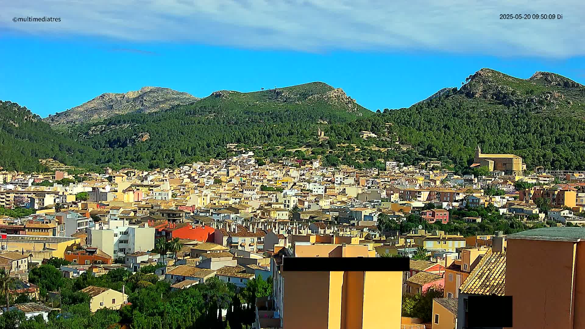 A sprawling town of low-rise buildings nestled in a valley, surrounded by lush green hills under a mostly clear blue sky.