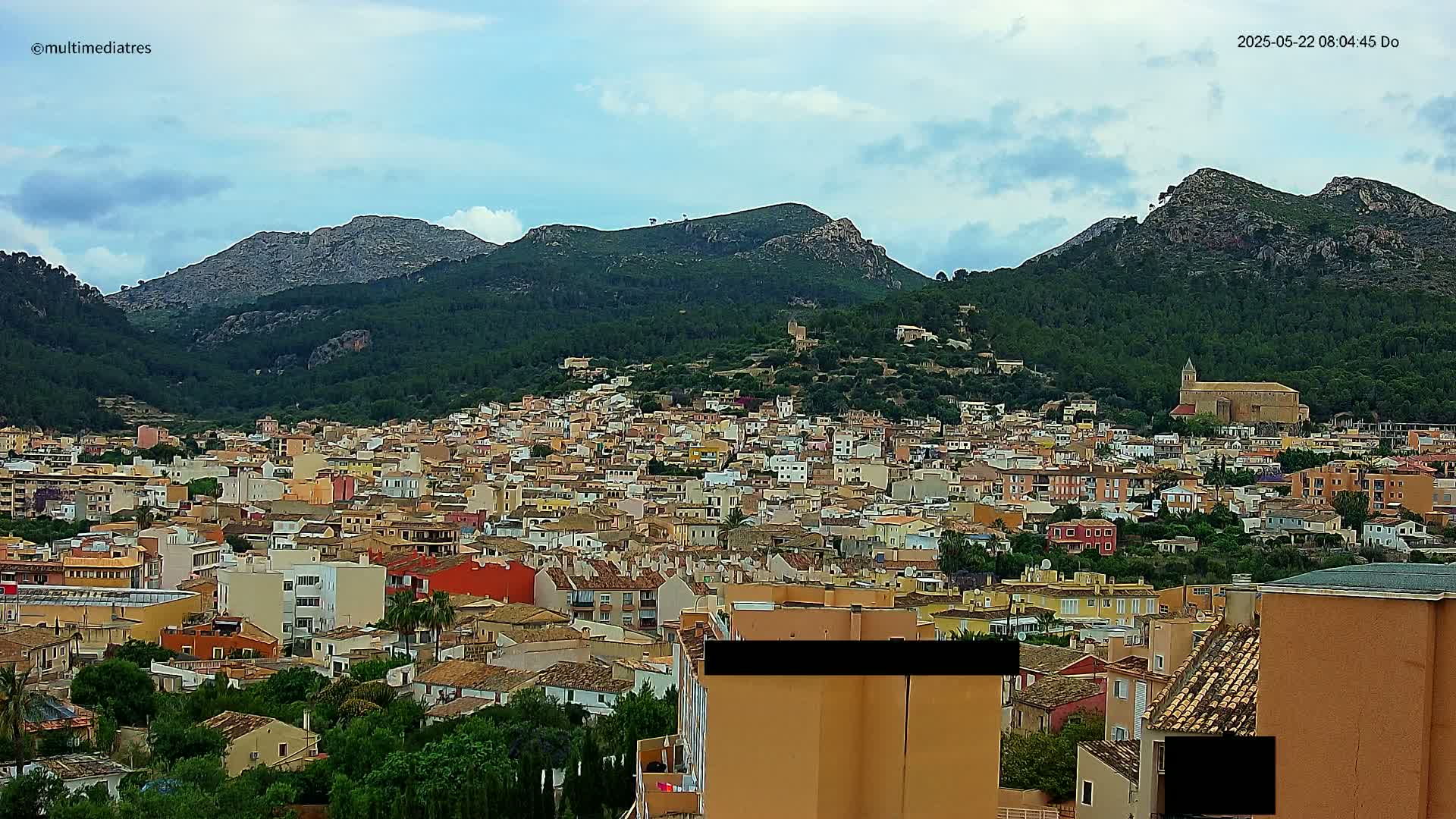 A sprawling town of low-rise buildings nestles at the foot of verdant, mountainous terrain under a partly cloudy sky.