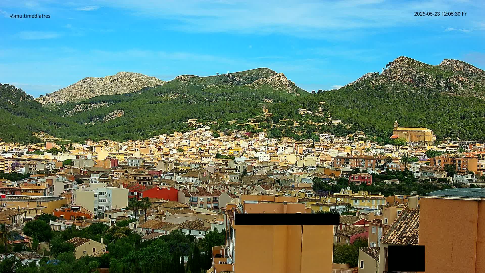 A dense cluster of buildings, predominantly low-rise and light-colored, stretches across a hillside, nestled beneath mountains under a clear blue sky.