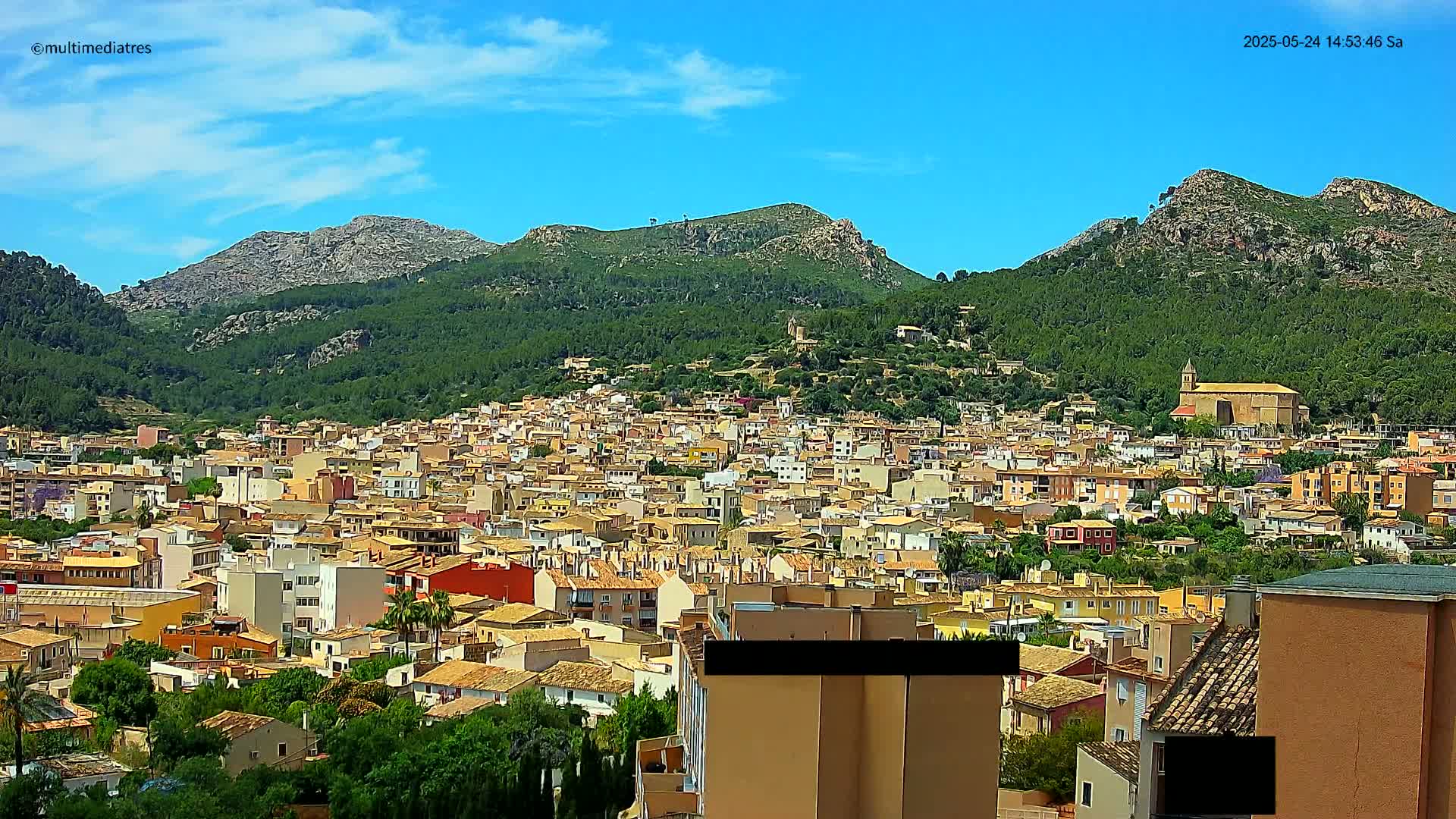 A sprawling town of tan and light brown buildings nestled in a valley surrounded by lush green mountains under a clear blue sky.