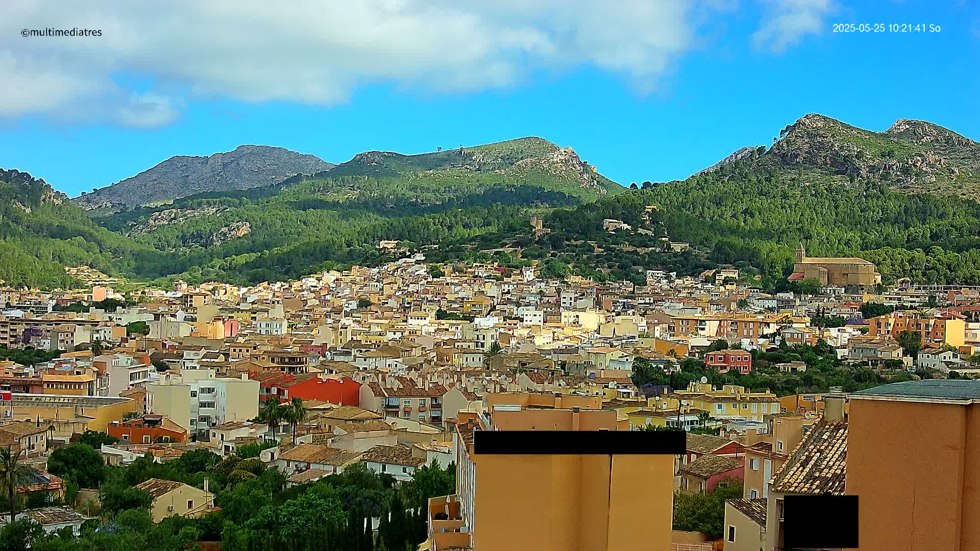 A sprawling town of low-rise buildings nestles in a valley surrounded by lush green mountains under a mostly clear blue sky.