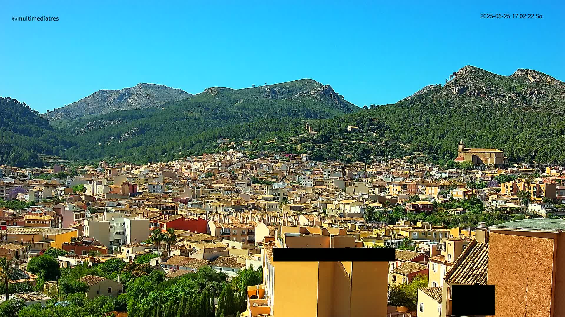 A sprawling town of low-rise buildings nestles at the foot of verdant, rocky mountains under a clear blue sky.