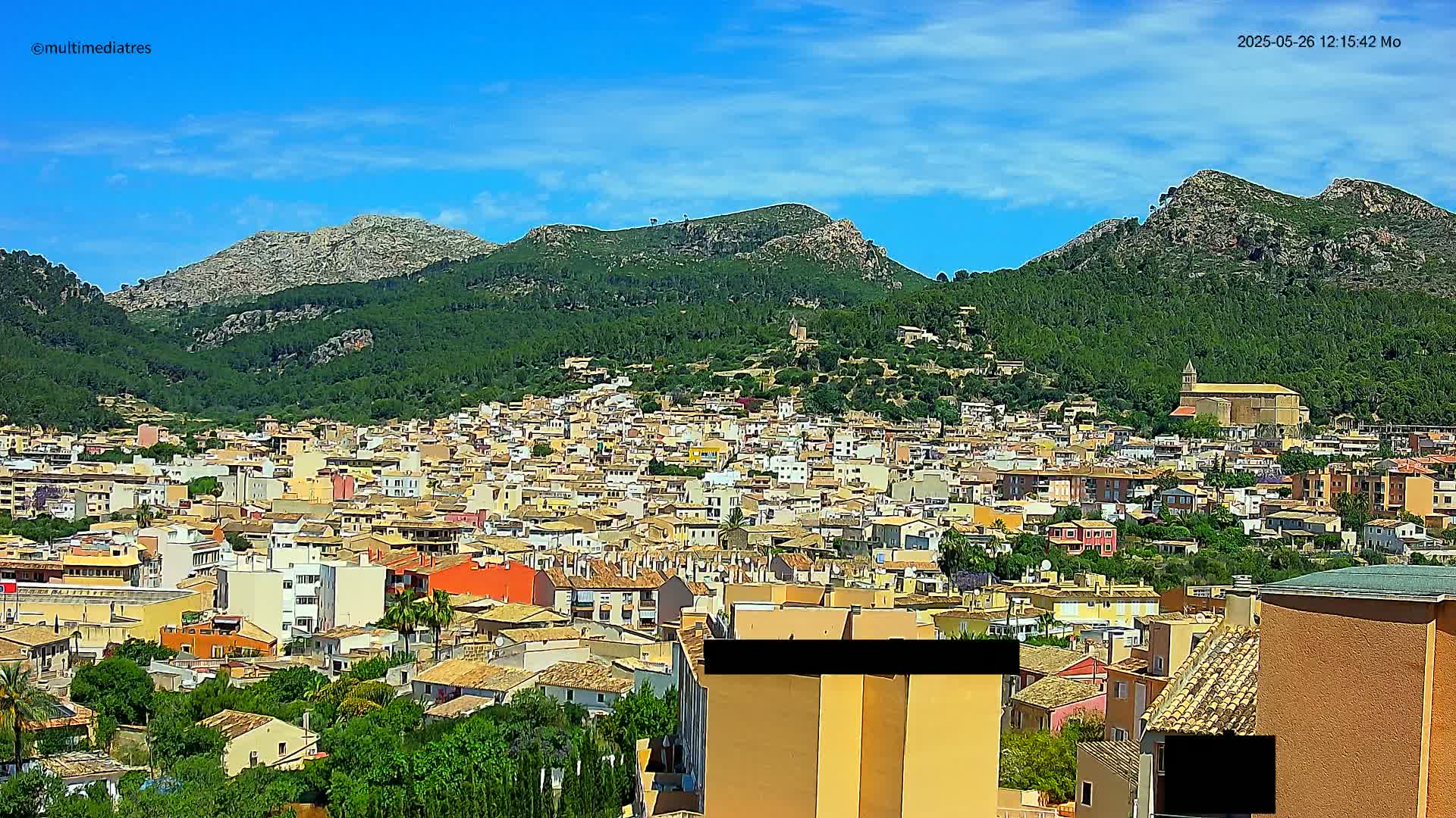 A sprawling town of closely packed, low-rise buildings nestles at the base of a lush, green mountain range under a bright, clear blue sky.