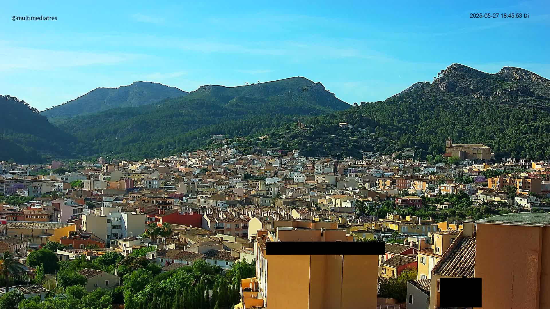 A sprawling town of low-rise buildings nestled in a valley, surrounded by lush green hills under a clear blue sky.