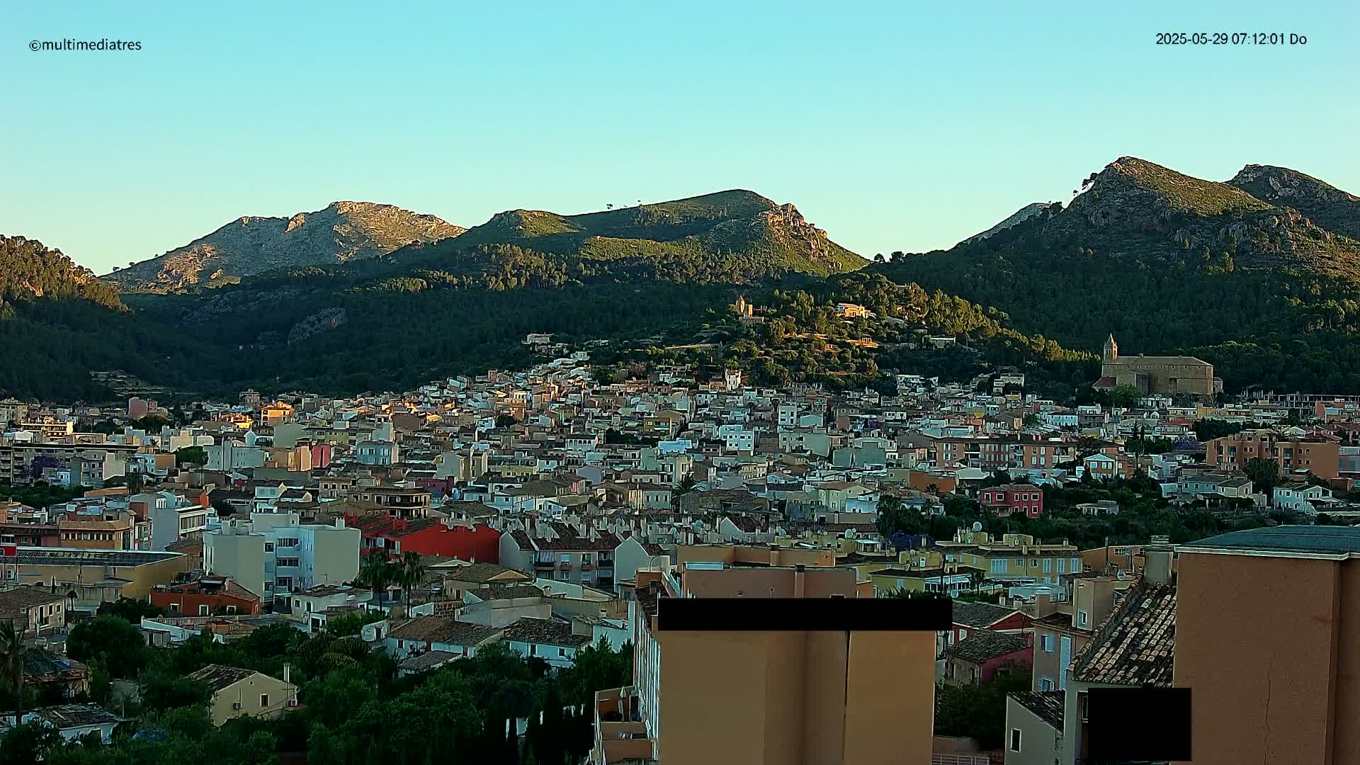 A sprawling town nestled in a valley between verdant, mountainous hills under a clear, bright sky.