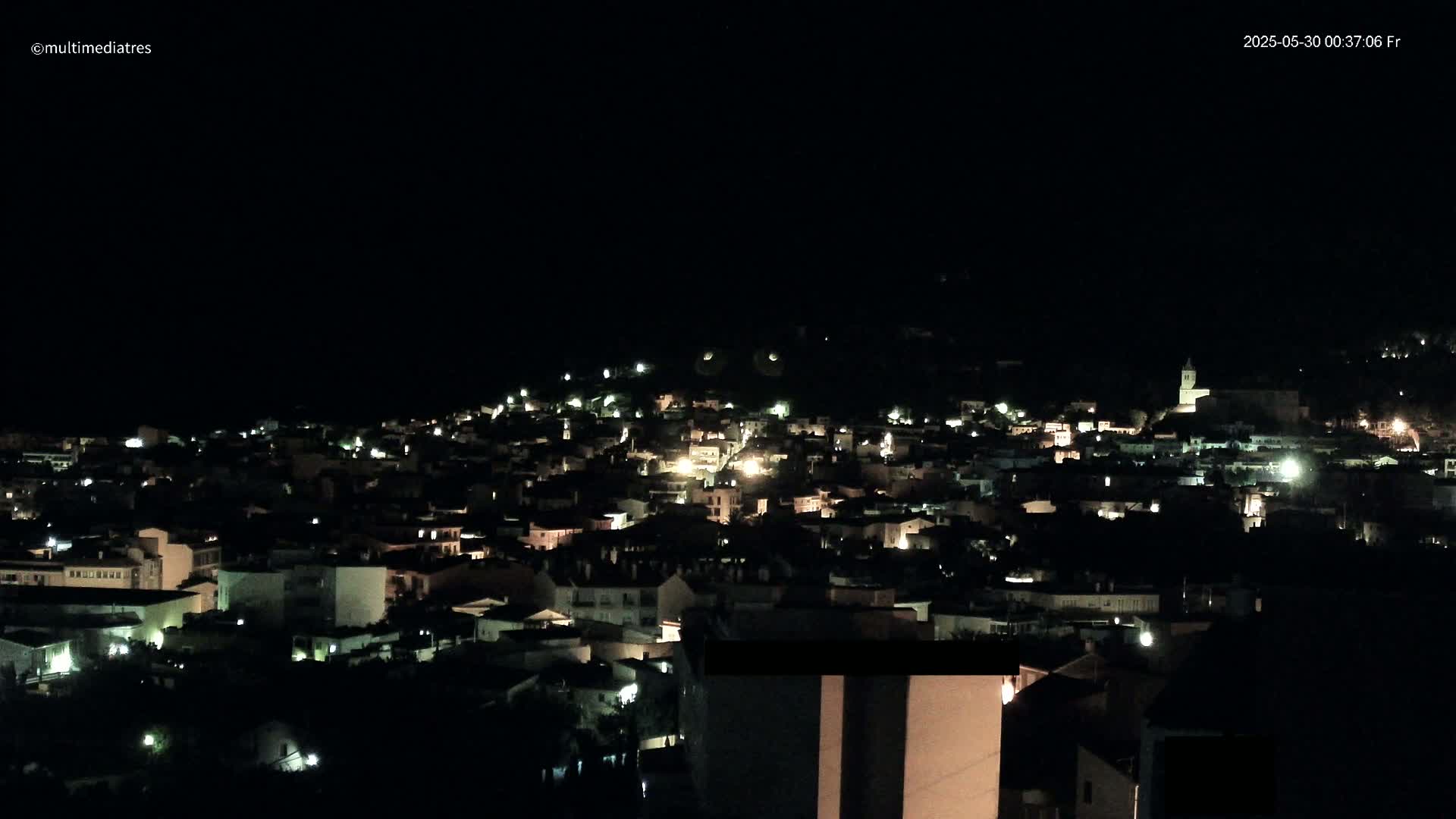 A nighttime, aerial view shows a small town nestled on a hill, its buildings illuminated by scattered lights under a clear, dark sky.