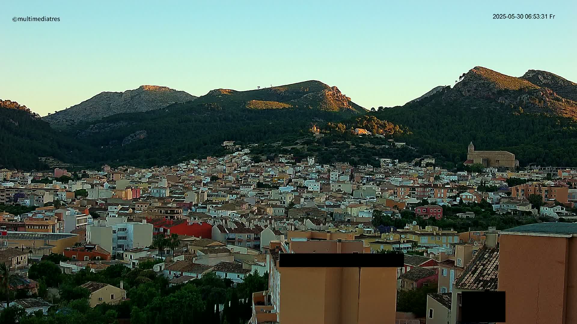 A sprawling town nestled at the base of sunlit, verdant mountains under a clear, pale-blue sky.