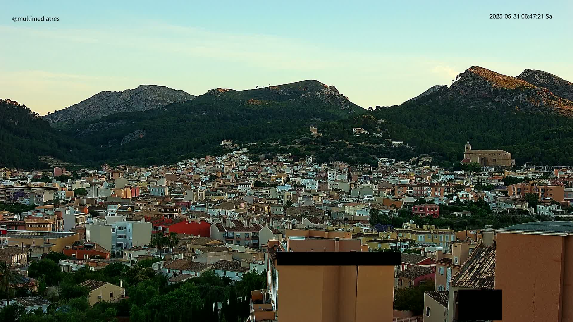 A sprawling town of low-rise buildings nestles at the base of several verdant, rocky hills under a clear, light-blue sky.