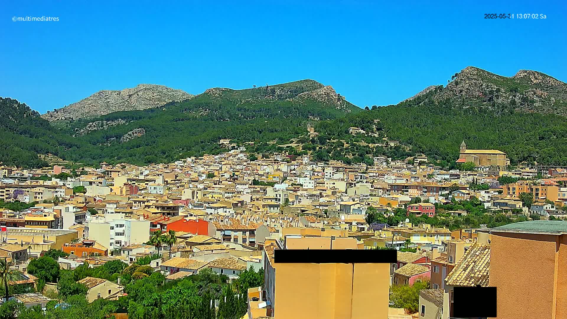 A sprawling town of tan and red-roofed buildings nestled at the base of lush green mountains under a clear blue sky.
