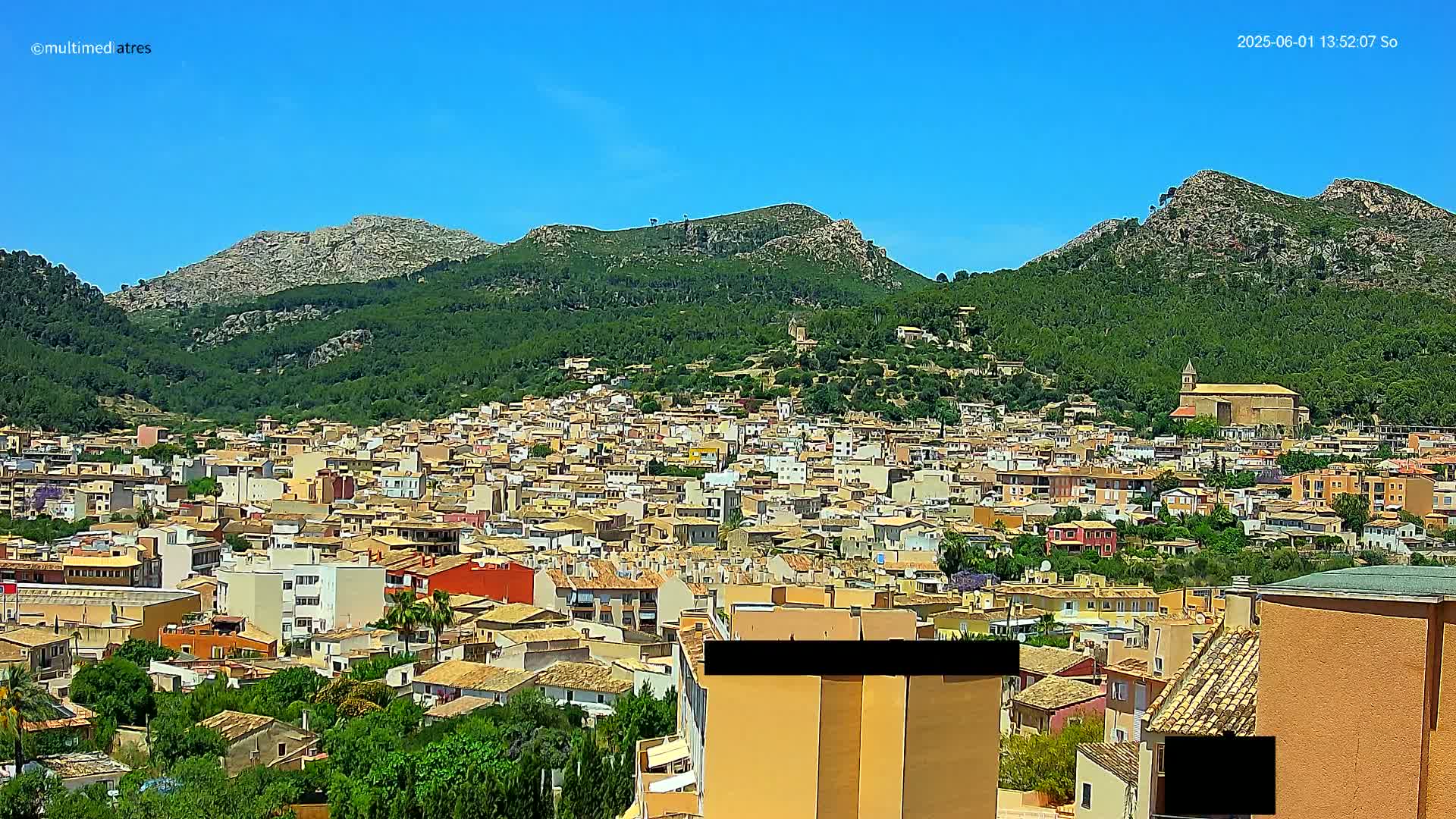 A sprawling town of tan and red-roofed buildings nestled at the base of a verdant mountain range under a clear blue sky.