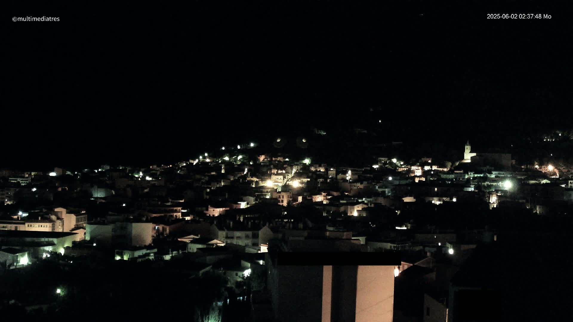 A nighttime, aerial view shows a small town illuminated by scattered lights on a hill under a clear, dark sky.