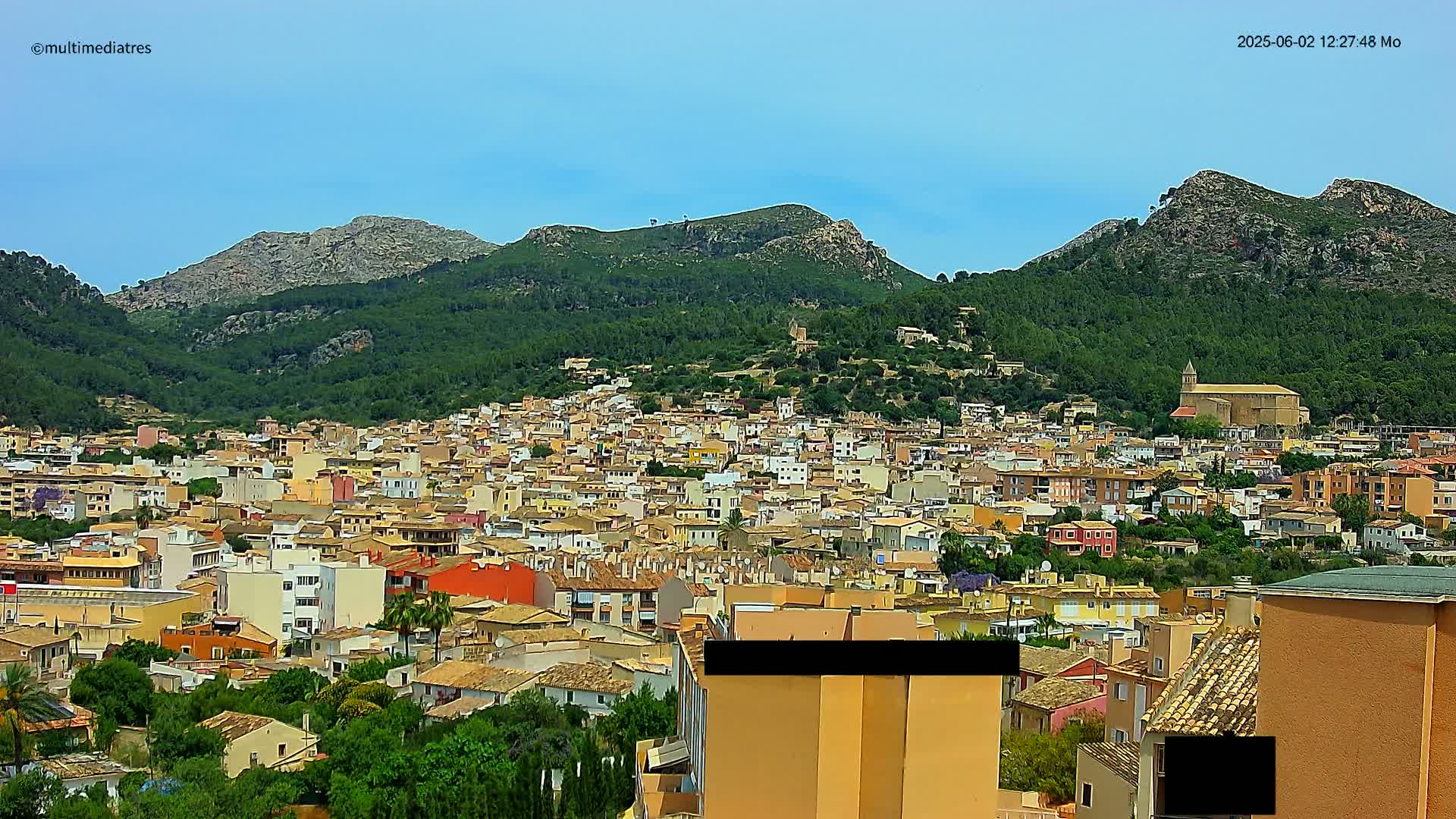 A dense collection of low-rise buildings, nestled at the base of lush green mountains under a clear blue sky.