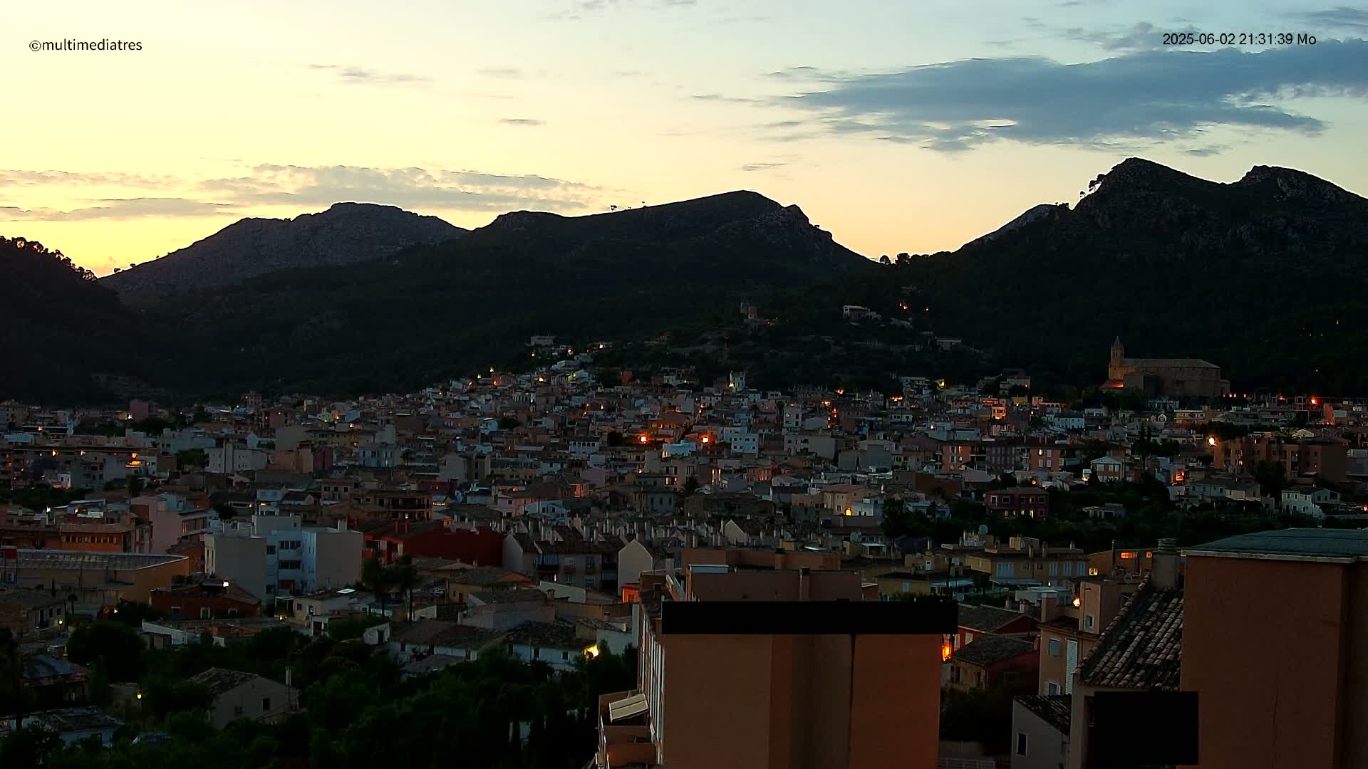 A town with many lights nestled at the base of dark, mountainous terrain under a twilight sky.