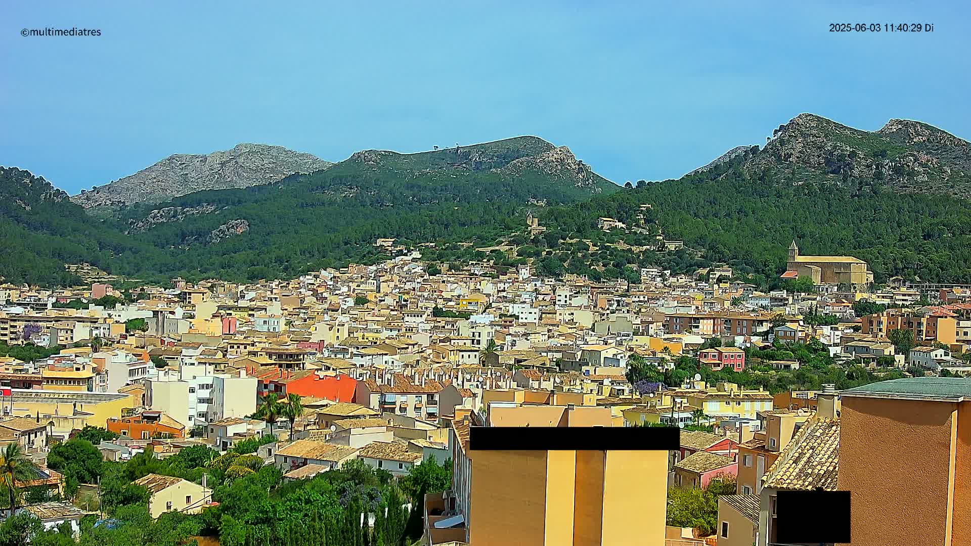 A dense collection of buildings, predominantly low-rise and light-colored, stretches across a hillside, backed by verdant mountains under a clear, bright sky.