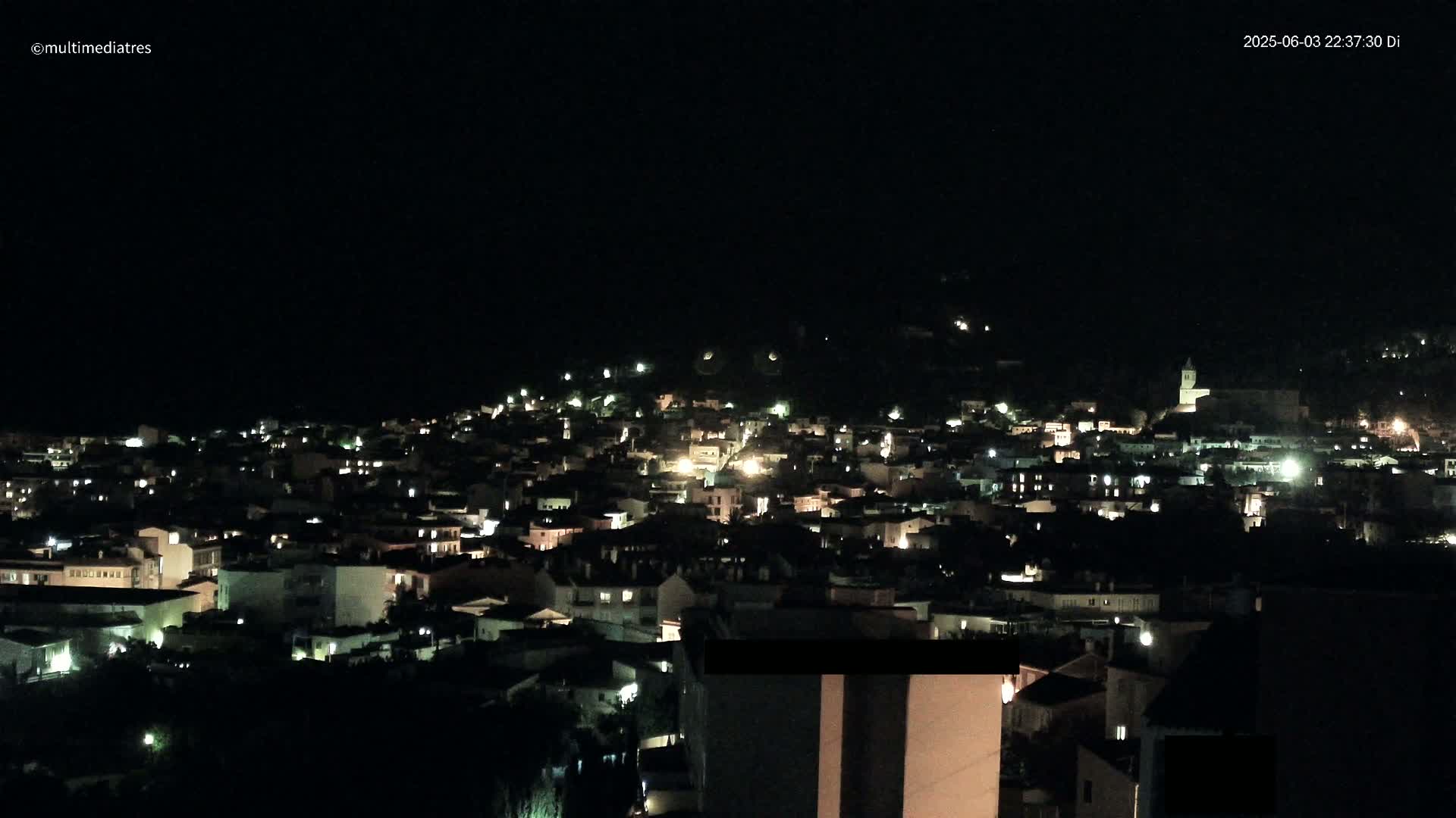 A nighttime view of a town nestled on a hillside, with scattered lights illuminating buildings under a clear, dark sky.