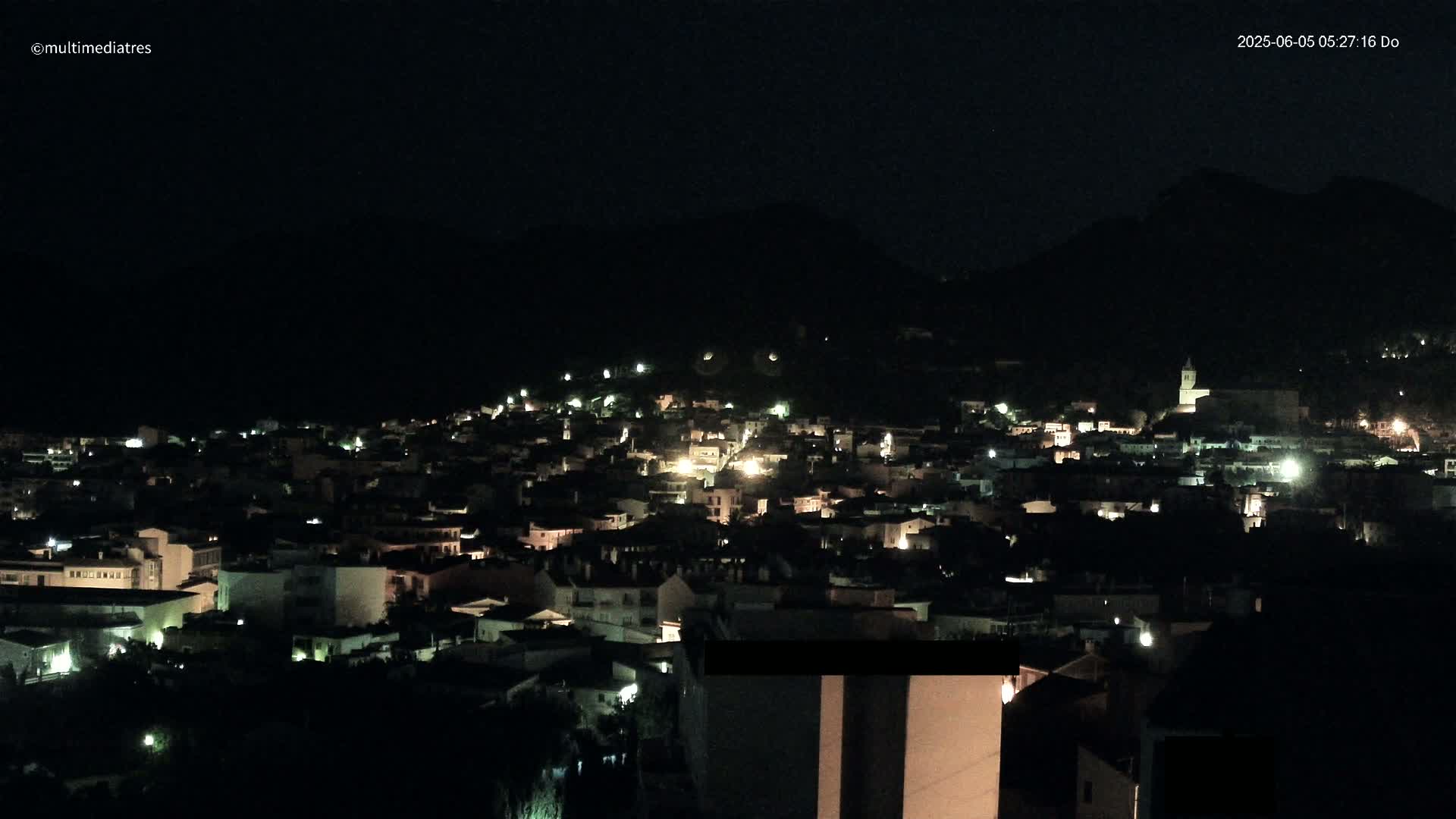 A nighttime view of a town nestled in a dark, hilly landscape, illuminated by scattered lights.