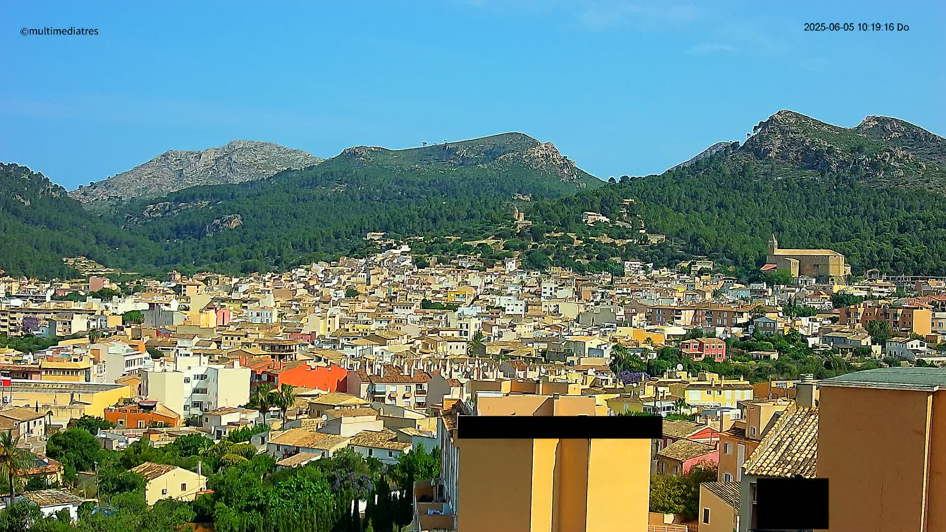 A sprawling town of closely packed buildings nestles in a valley surrounded by lush green hills and rocky mountains under a clear blue sky.