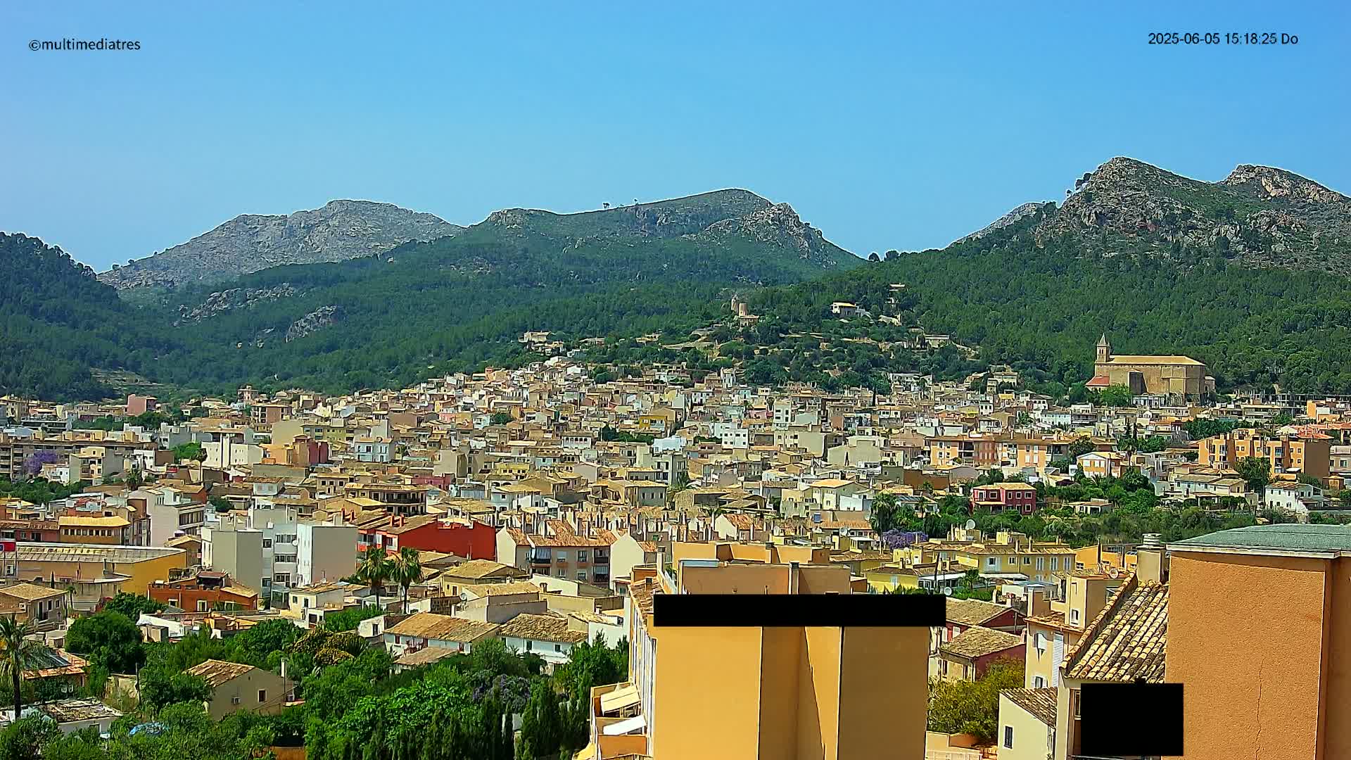 A sprawling town of low-rise buildings nestles at the foot of verdant, rocky mountains under a clear blue sky.