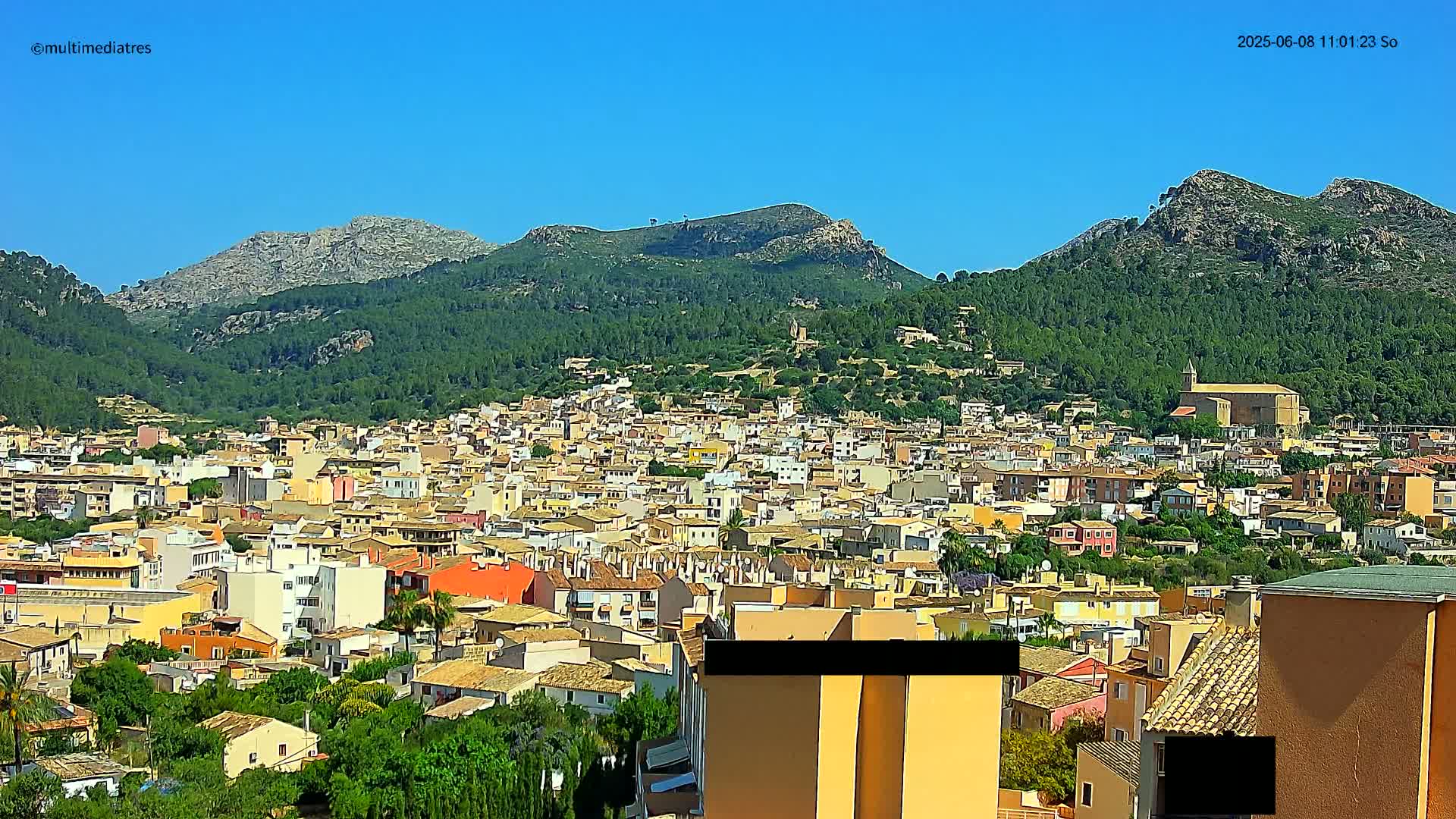 A sprawling town of low-rise buildings nestled in a valley, surrounded by lush green hills and mountains under a clear blue sky.