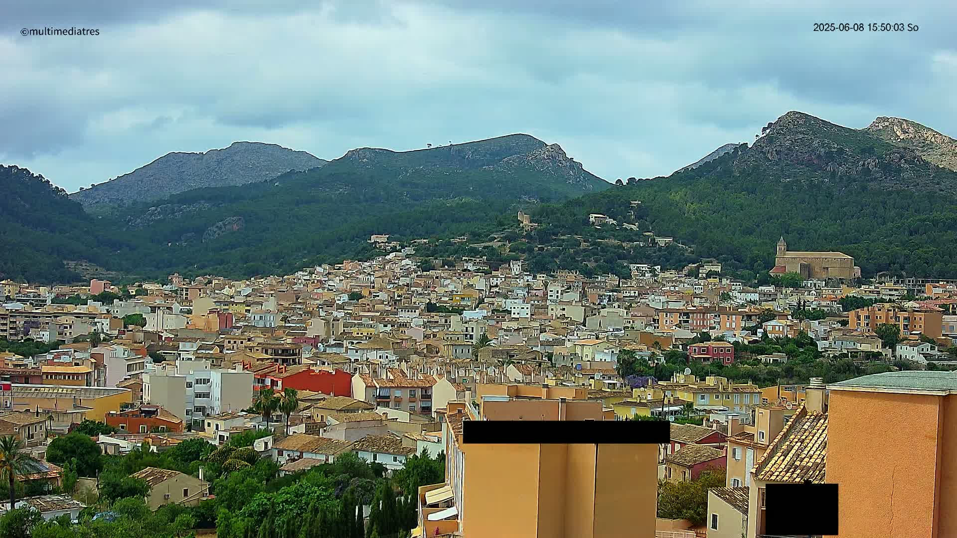 A sprawling town of low-rise buildings nestles at the foot of verdant, rocky mountains under a cloudy sky.