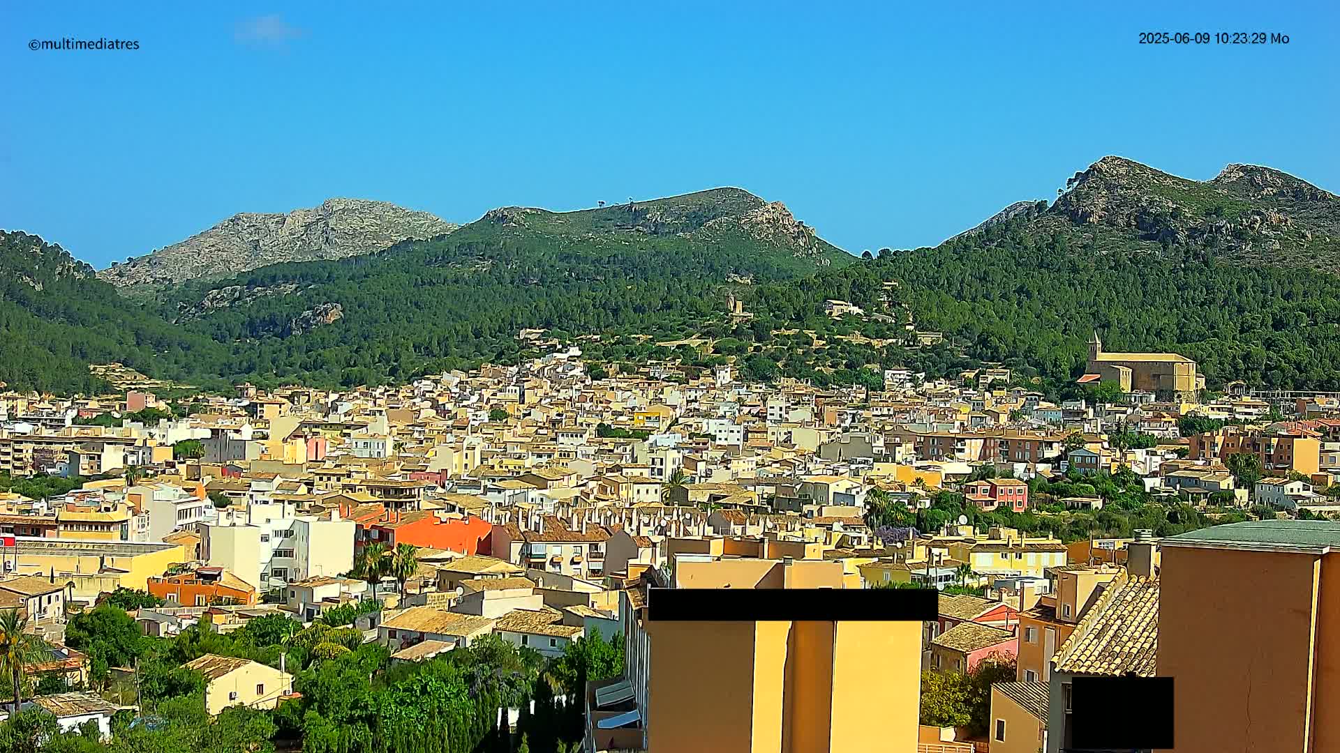 A sprawling town of low-rise buildings nestles in a verdant valley, backed by several rocky mountains under a clear blue sky.