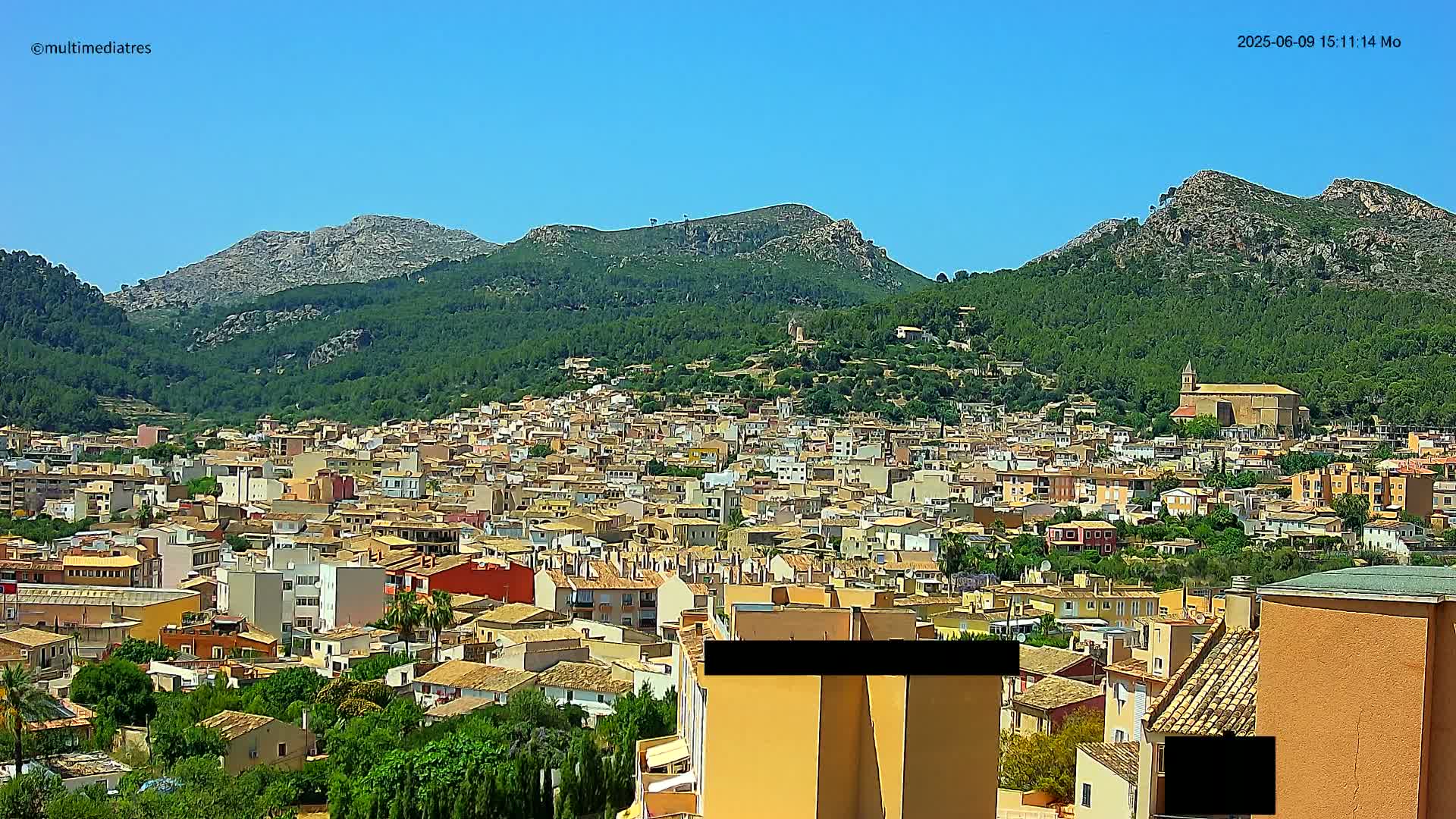 A sprawling town of tan and red-roofed buildings nestled in a verdant valley, backed by steep, rocky mountains under a clear blue sky.