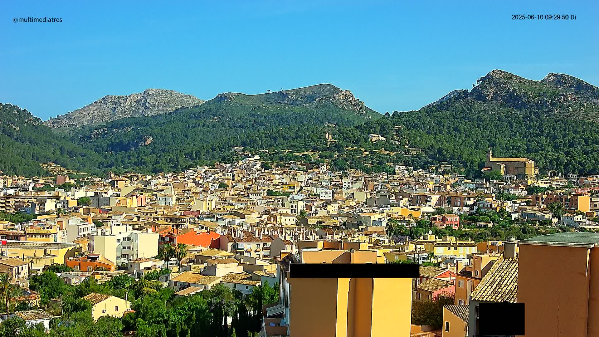 A sprawling town of tan and orange buildings nestles in a valley surrounded by lush green hills and mountains under a clear blue sky.