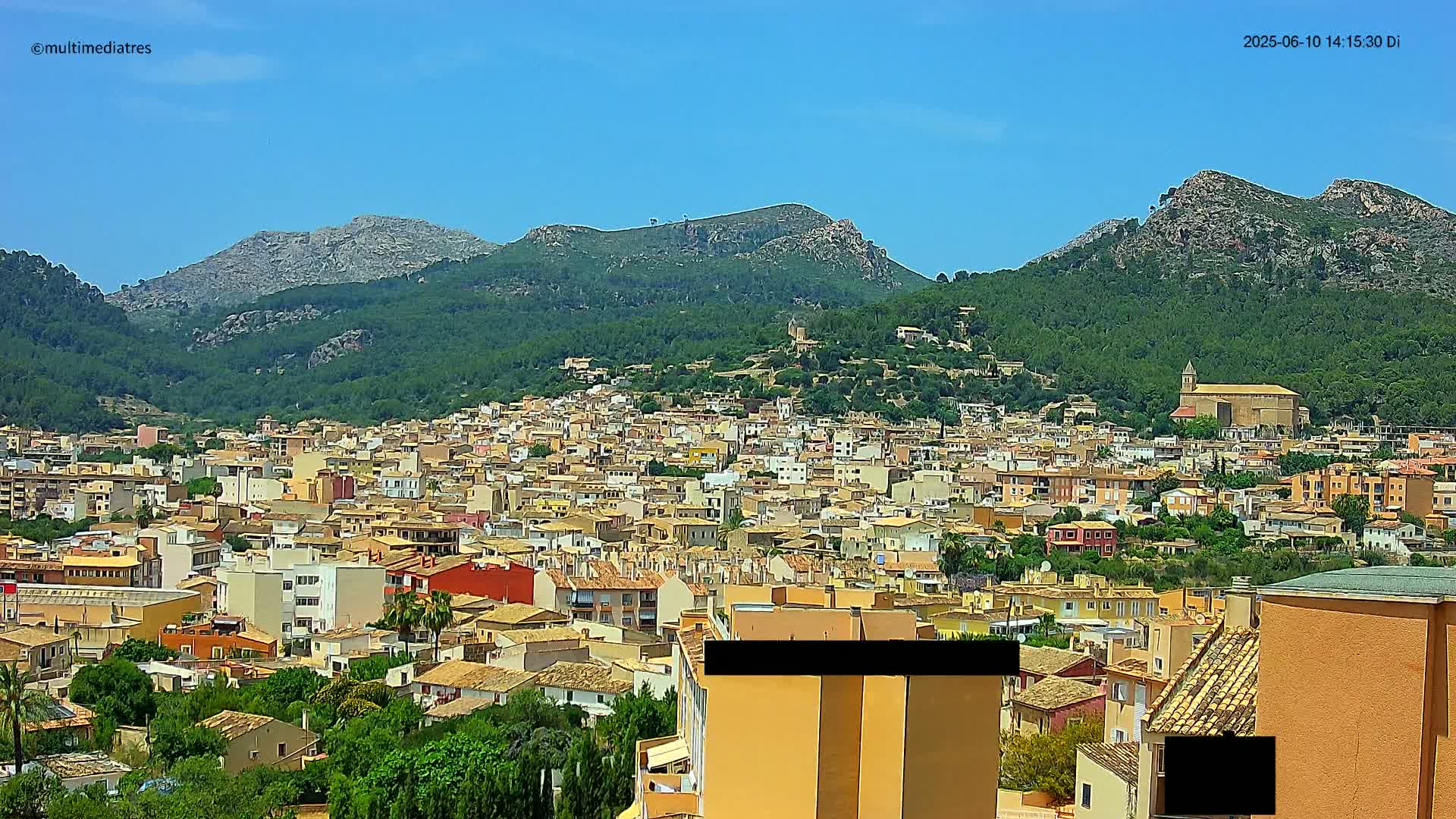 A sprawling town of low-rise buildings nestled in a valley, surrounded by lush green hills and mountains under a clear blue sky.