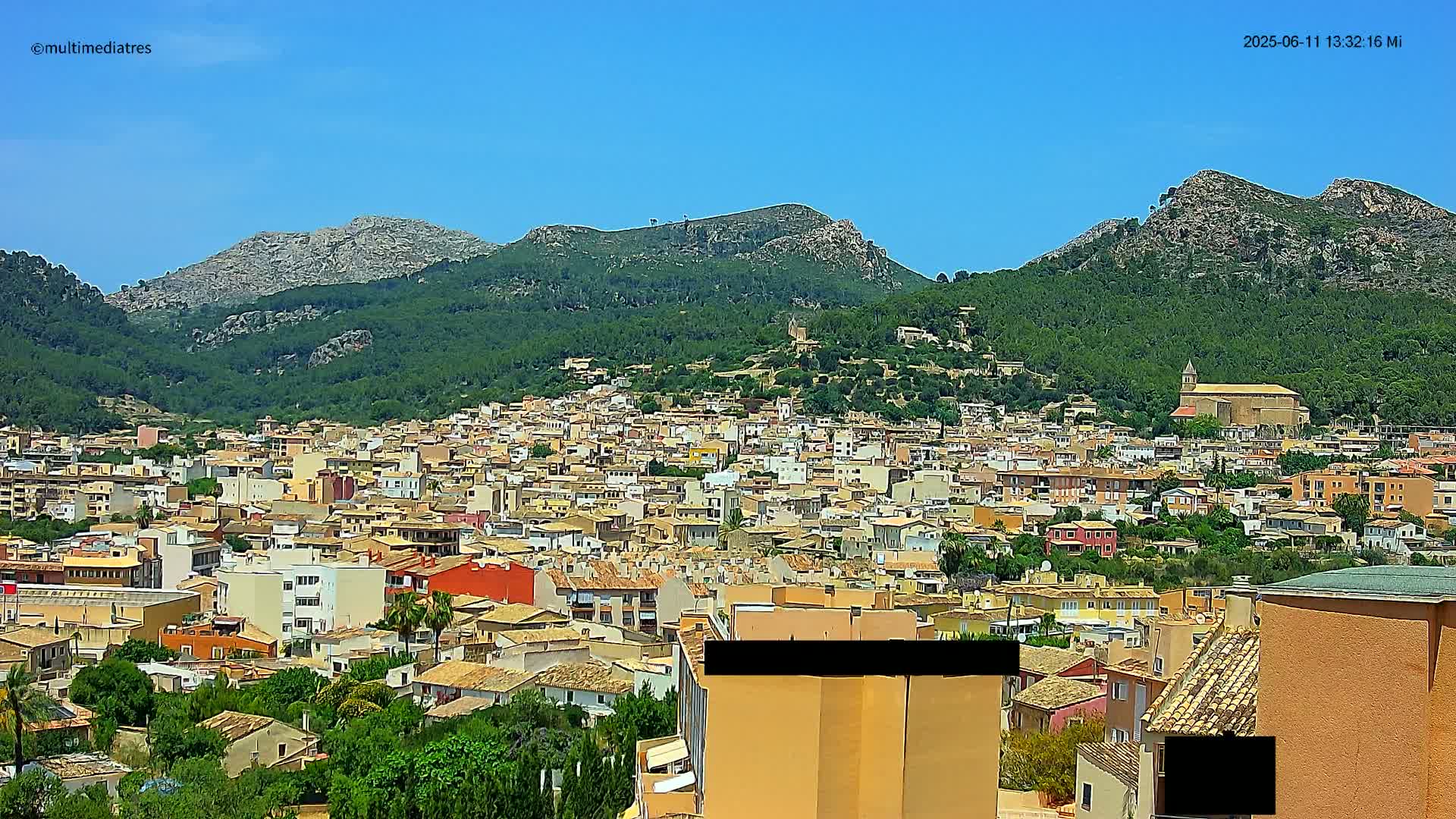 A sprawling town of tan and orange buildings nestled in a verdant valley, backed by rocky, tree-covered mountains under a clear blue sky.