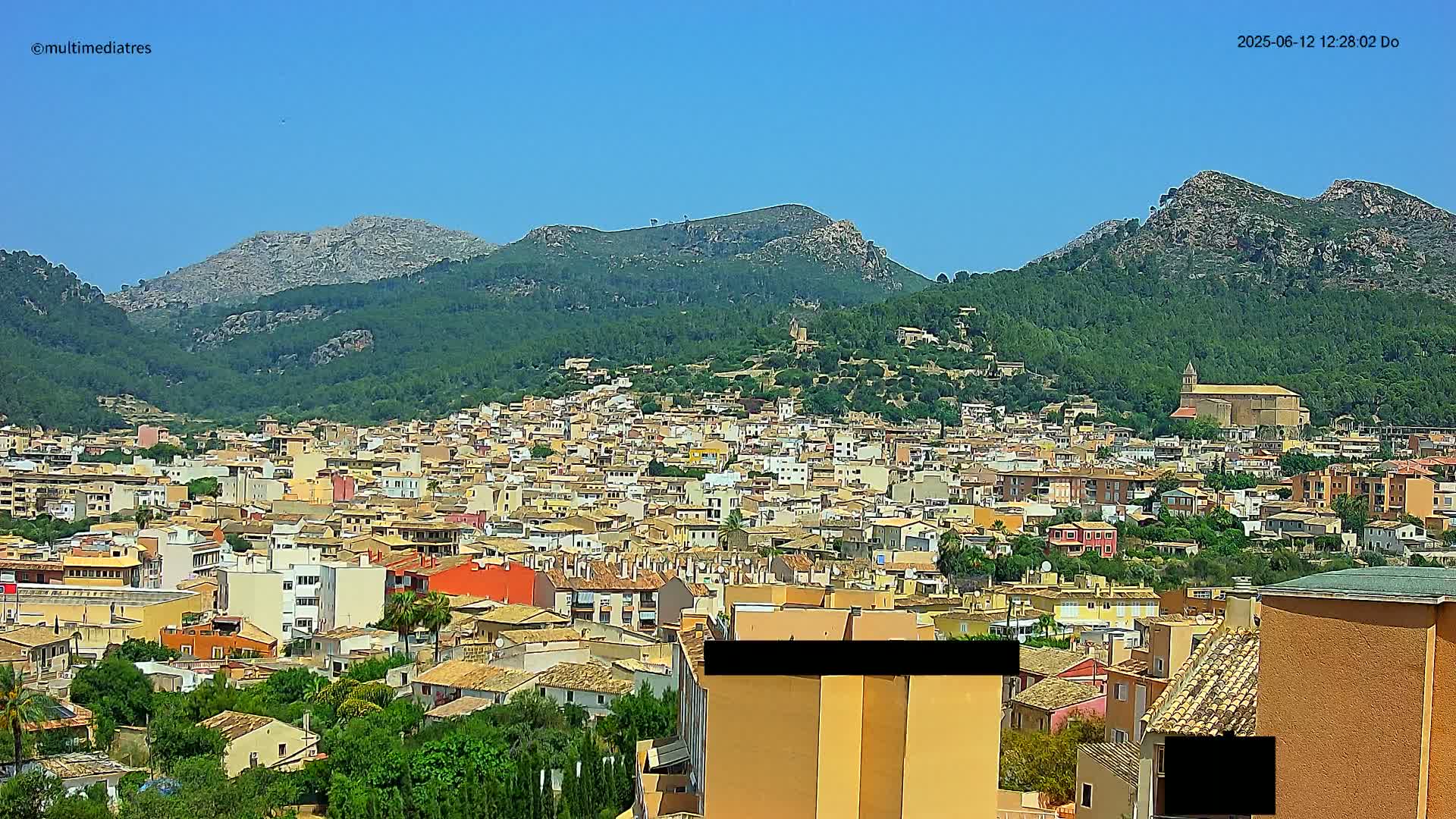 A sprawling town nestled at the foot of verdant, rocky mountains under a clear blue sky.
