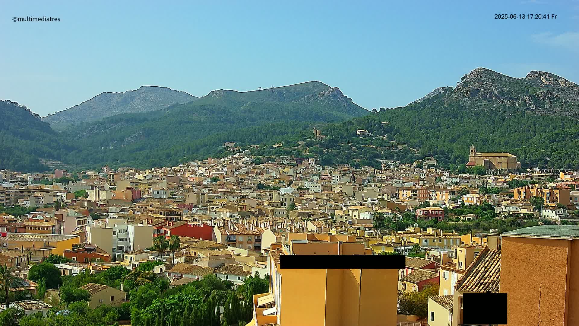 A sprawling town of low-rise buildings nestled in a valley between verdant, mountainous terrain under a clear, sunny sky.