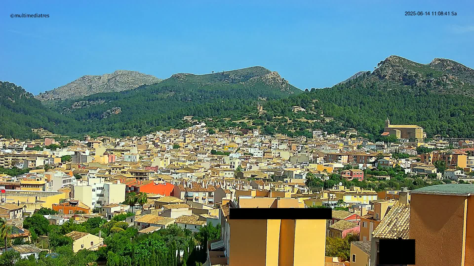 A sprawling town of closely packed buildings nestles at the base of lush green hills under a clear blue sky.