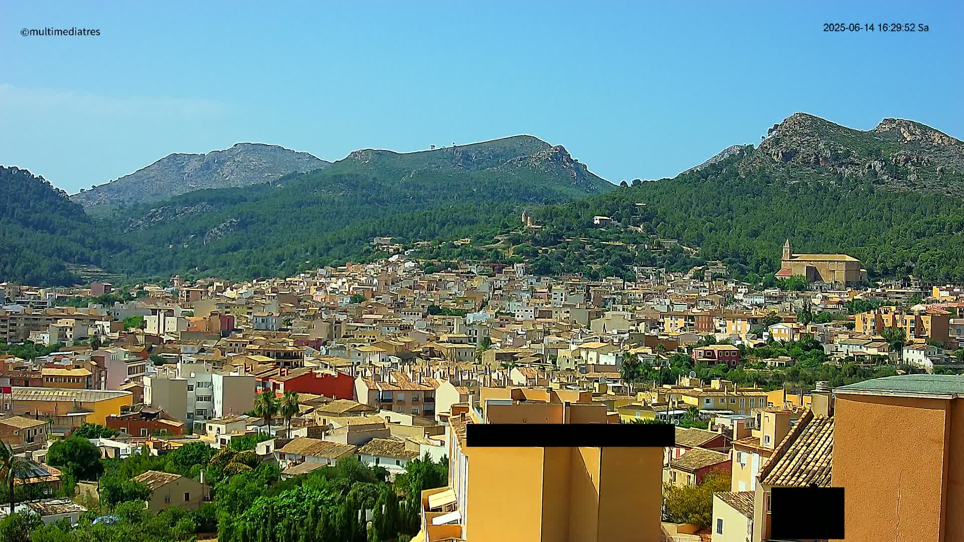 A sprawling town of low-rise buildings nestles against a backdrop of verdant, mountainous terrain under a clear blue sky.
