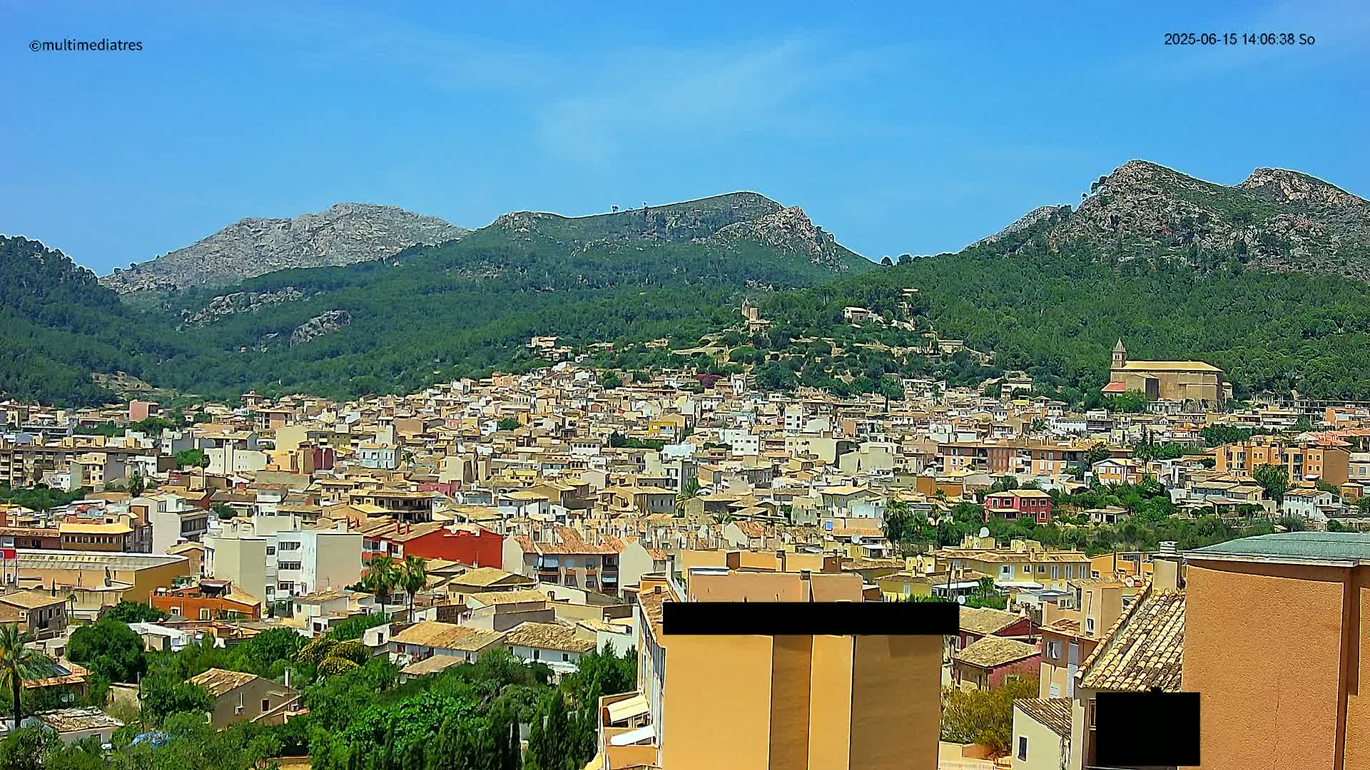 A sprawling town of tan and red-roofed buildings nestled in a verdant valley, backed by rocky, tree-covered mountains under a clear blue sky.