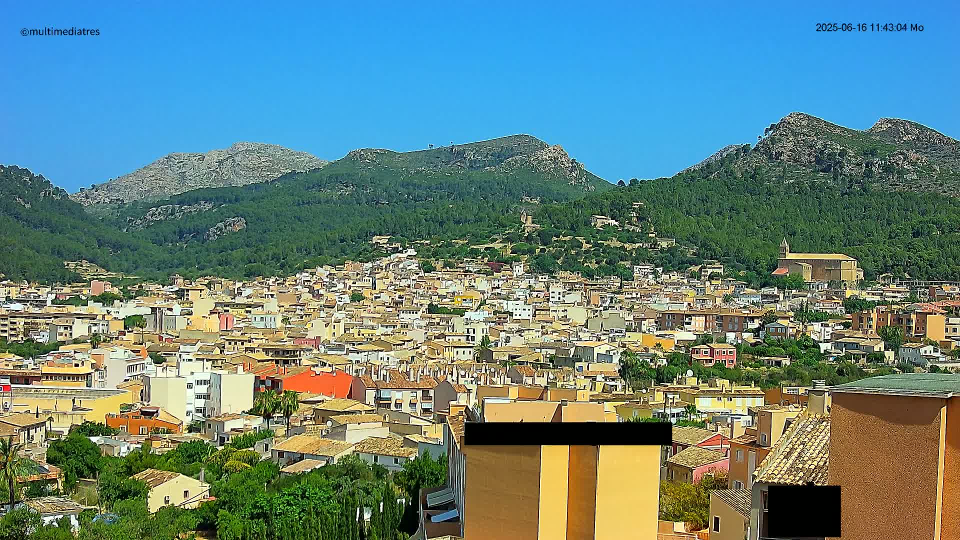 A sprawling town of closely packed buildings nestles in a valley surrounded by lush green hills and mountains under a clear blue sky.