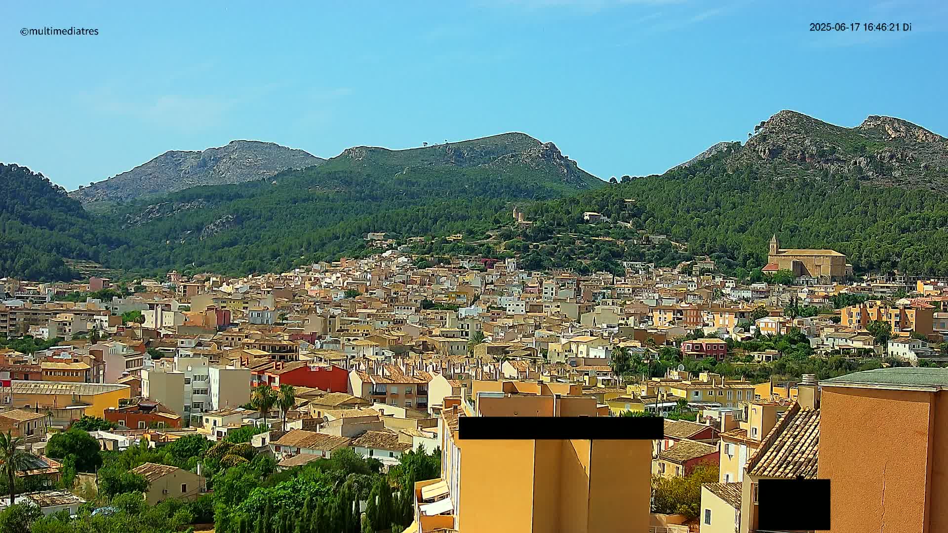 A sprawling town of low-rise buildings nestles in a valley surrounded by lush green mountains under a clear blue sky.