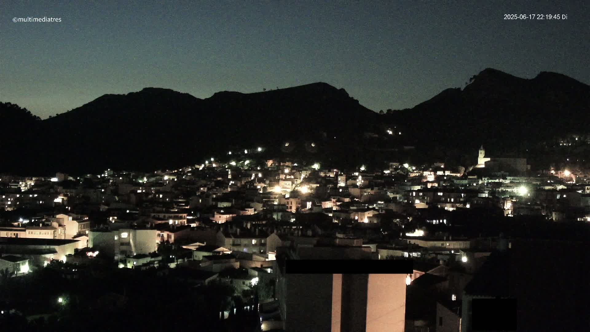 A nighttime view of a town nestled against a dark mountain under a clear, dark sky.